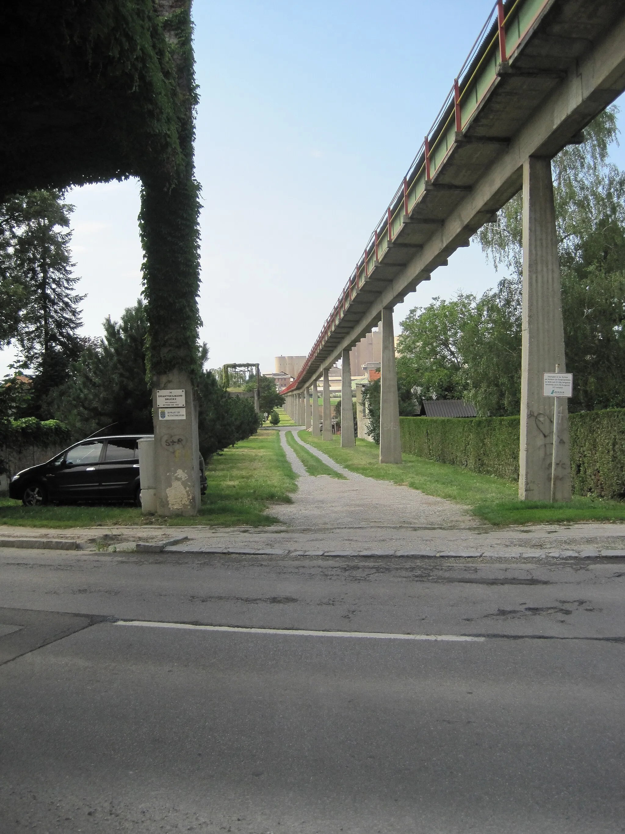 Photo showing: A conveyor device that transports limestone from quarry to factory, passing through the town of Mannersdorf am Leithagebirge in Austria.  On the left are the supports for a former funicular system that served the same function until 1960.  In the background is the Lafarge-Perlmooser plant fed by the system.