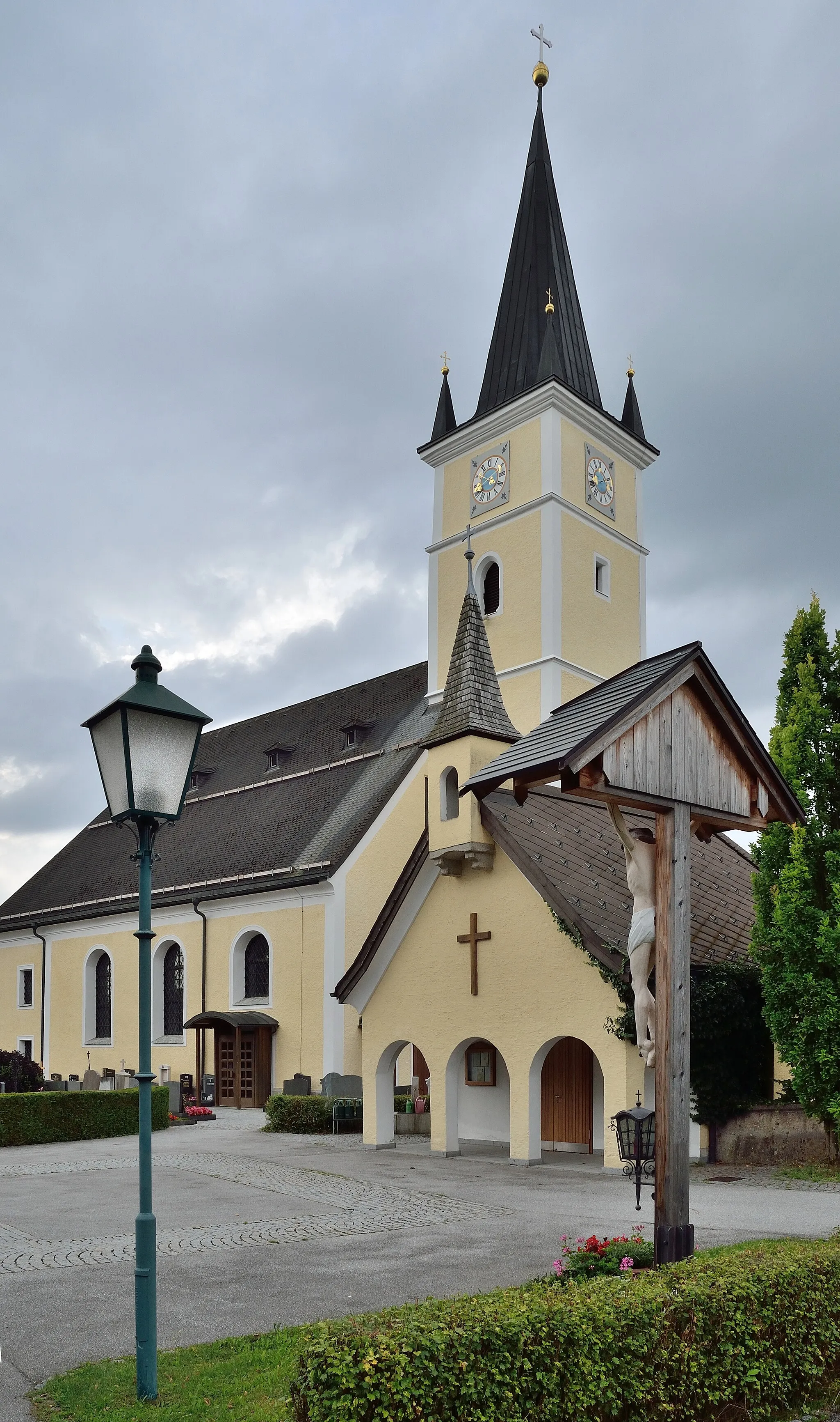 Photo showing: Der Friedhof Henndorf umgibt die Pfarrkirche. Hier das Friedhofskreuz im neueren Teil, die Aufbahrungshalle und die Pfarrkirche im Hintergrund.