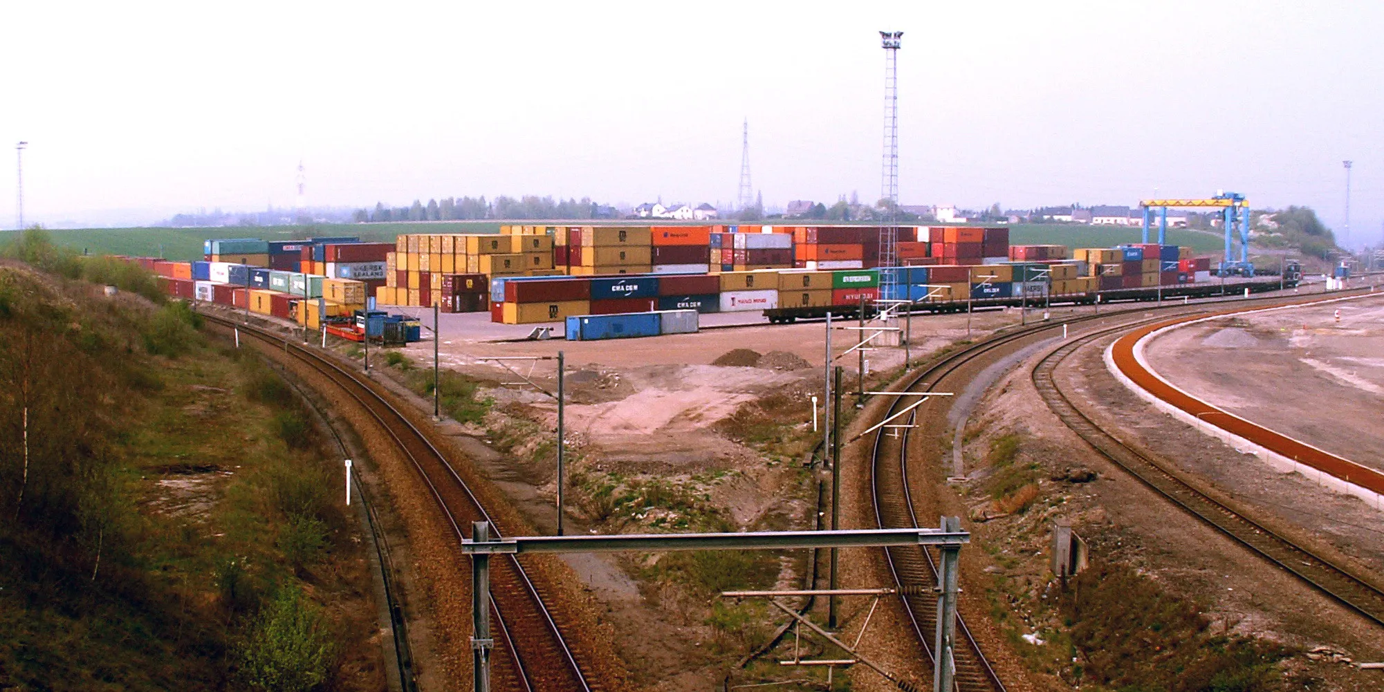 Photo showing: The container Terminal of Athus, as seen from Road N830. The terminal is divided by railway line 167 to Arlon (on the right) and bordered by line 165/1 to Virton (on the left) and line 165 (behind the stacks). Both coming form the Luxembourgian city of Rodange. Photo shows the western zone of the terminal. A second (eastern) zone is available, but out of the photograph. This configuration was visible between 2004 and 2019, and updated to allow a new branch (Line 165/3 to Mont-Saint Martin)