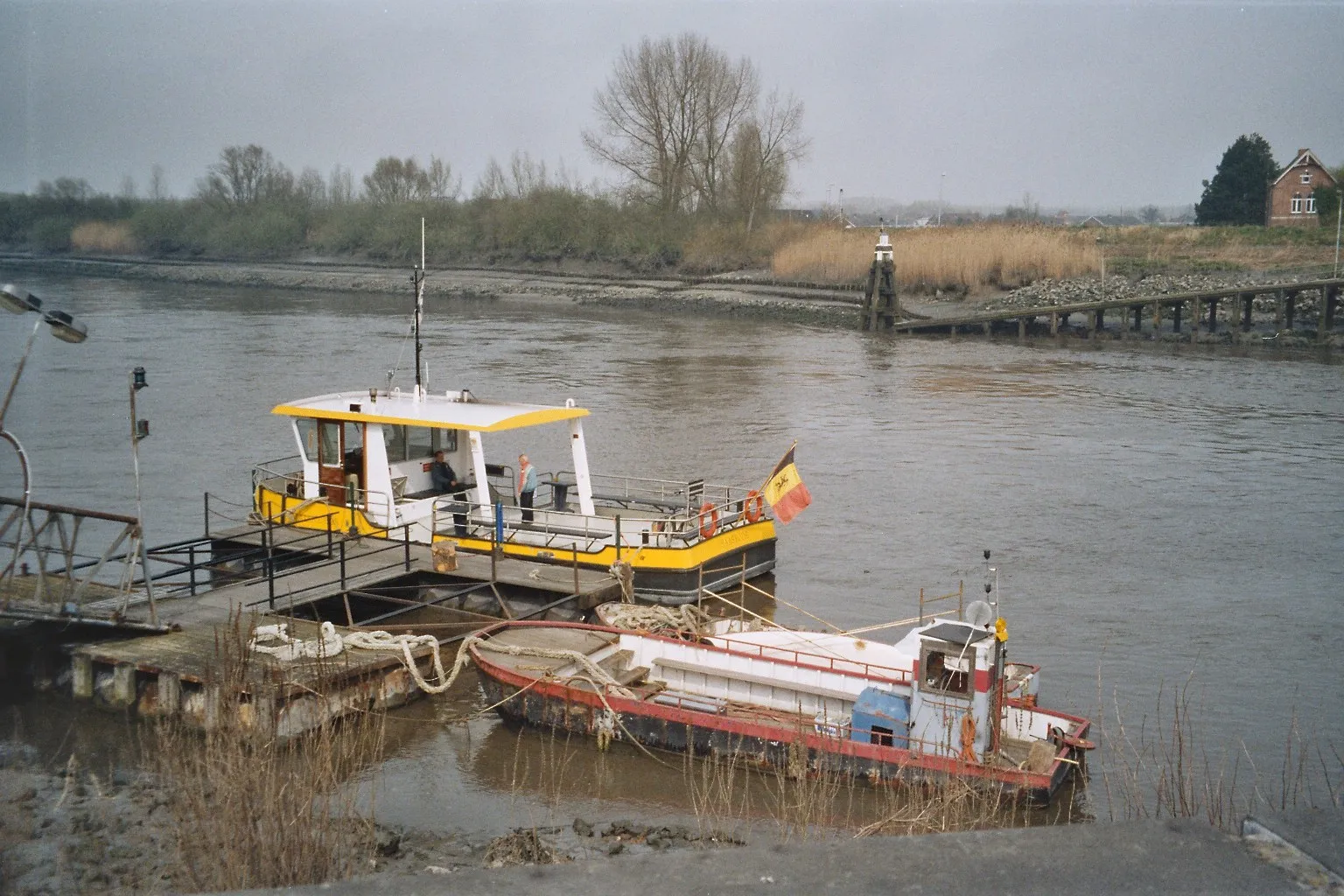 Photo showing: Two small (only for pedestrians and cyclists) ferries across Scheldt river in town Baasrode (Belgium), which is a part of Dendermonde munitipality. Previously, the small open boat (on the right) was used. Now it is replaced by bigger boat (on the photo the one with the flag)