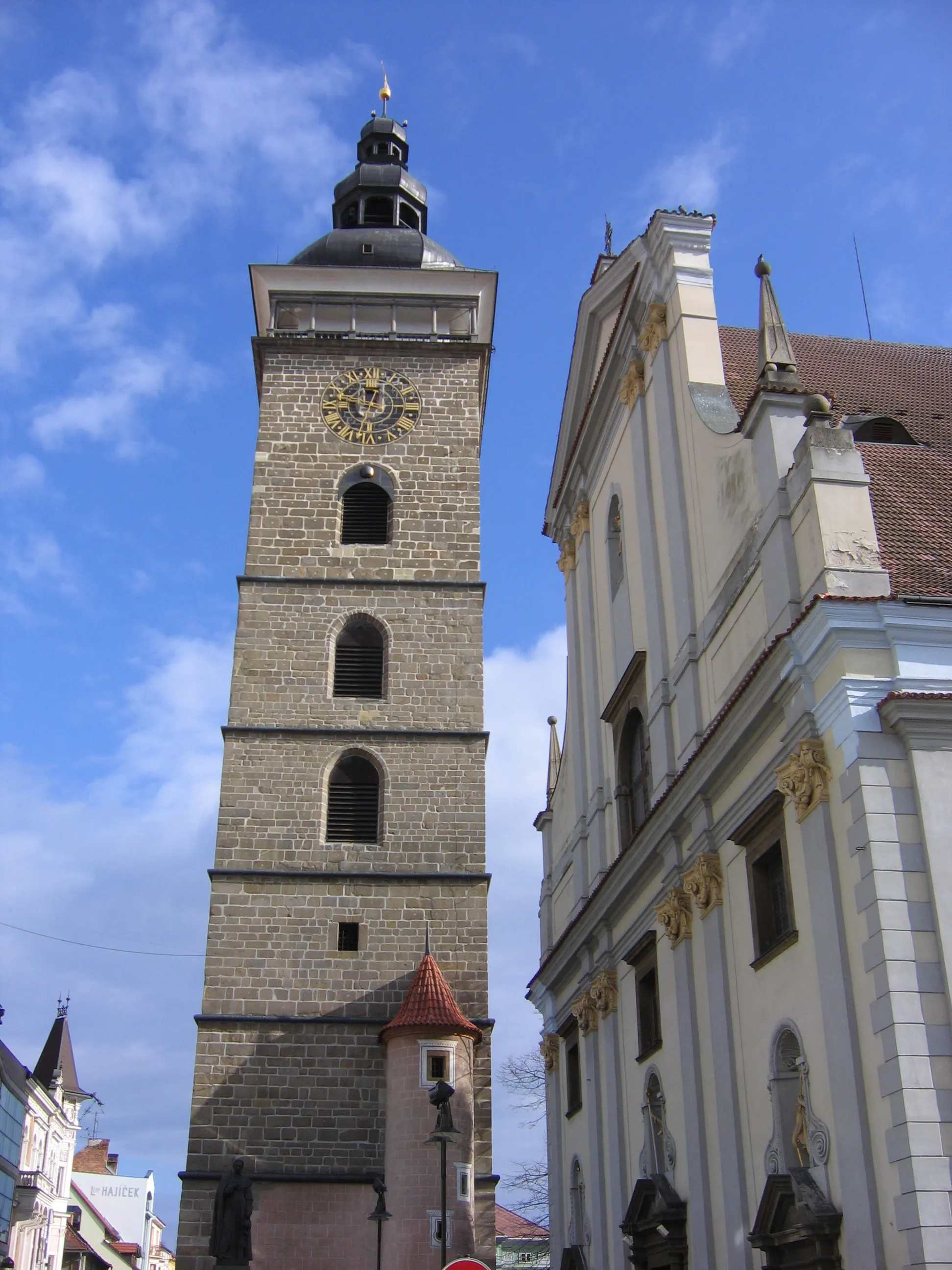 Photo showing: České Budějovice, Czech Republic: Black Tower with clock and bells, on right is cathedral of Sankt Nicolas. Before Black Tower is statue of Jan Valerián Jirsík, 4th bishop of České Budějovice