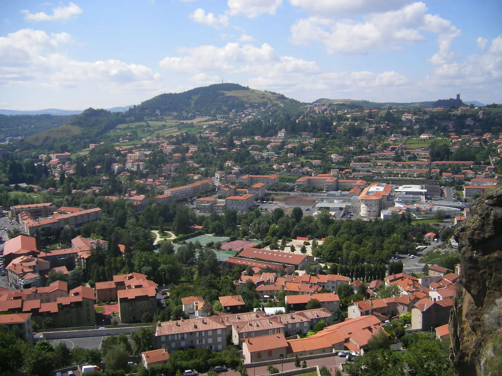 Photo showing: Le Puy-en-Velay, Aighuilhe, Polignac et Espaly-Saint-Marcel, Haute-Loire, Auvergne, France. Viewing west-north-west from the high-up perched Saint-Michel church of Aighuilhe.
At the forefront, the green squares are the tennis courts next to the camping site of Aiguilhe; but the two furthest courts are in the territory of Le Puy.
On the left near the edge of the photo, the set of buildings forming a near-square is Le Puy's fire station, along the place de la République.
The Borne river flows left to right (west-east) just behind the fire station and the camping site; a continuous ribbon of trees marks its course.
Facing the camping site on the opposite side of the river, is the Emile Roux hospital (on Le Puy-en-Velay) spreading from the right side of the photo over 2/3 of its width.
Mid-distance on the right: Polignac, with on the horizon line its recently rebuilt dungeon of the castle of Polignac. (Polignac reaches up to the left bank of the Borne opposite the fire station.)
Mid-distance on the left: Espaly-Saint-Marcel.

In the middle: the inactive volcano of Mont Denise with its antenna at the top and, to its left slightly behind, a volcanic rock formation known as Orgues d'Espaly.