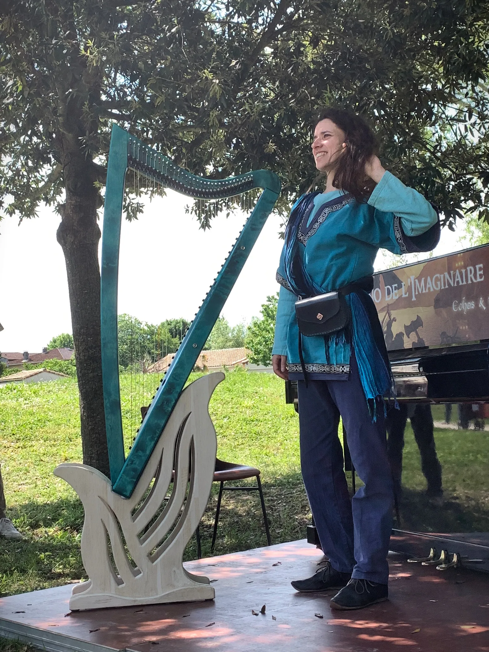 Photo showing: À gauche, sur un socle ouvragé en bois clair, une harpe celtique bleu turquoise. À droite, debout à coté de la harpe, la harpeuse et chanteuse Morgan of Glencoe en costume de scène bleu, souriant à son public.