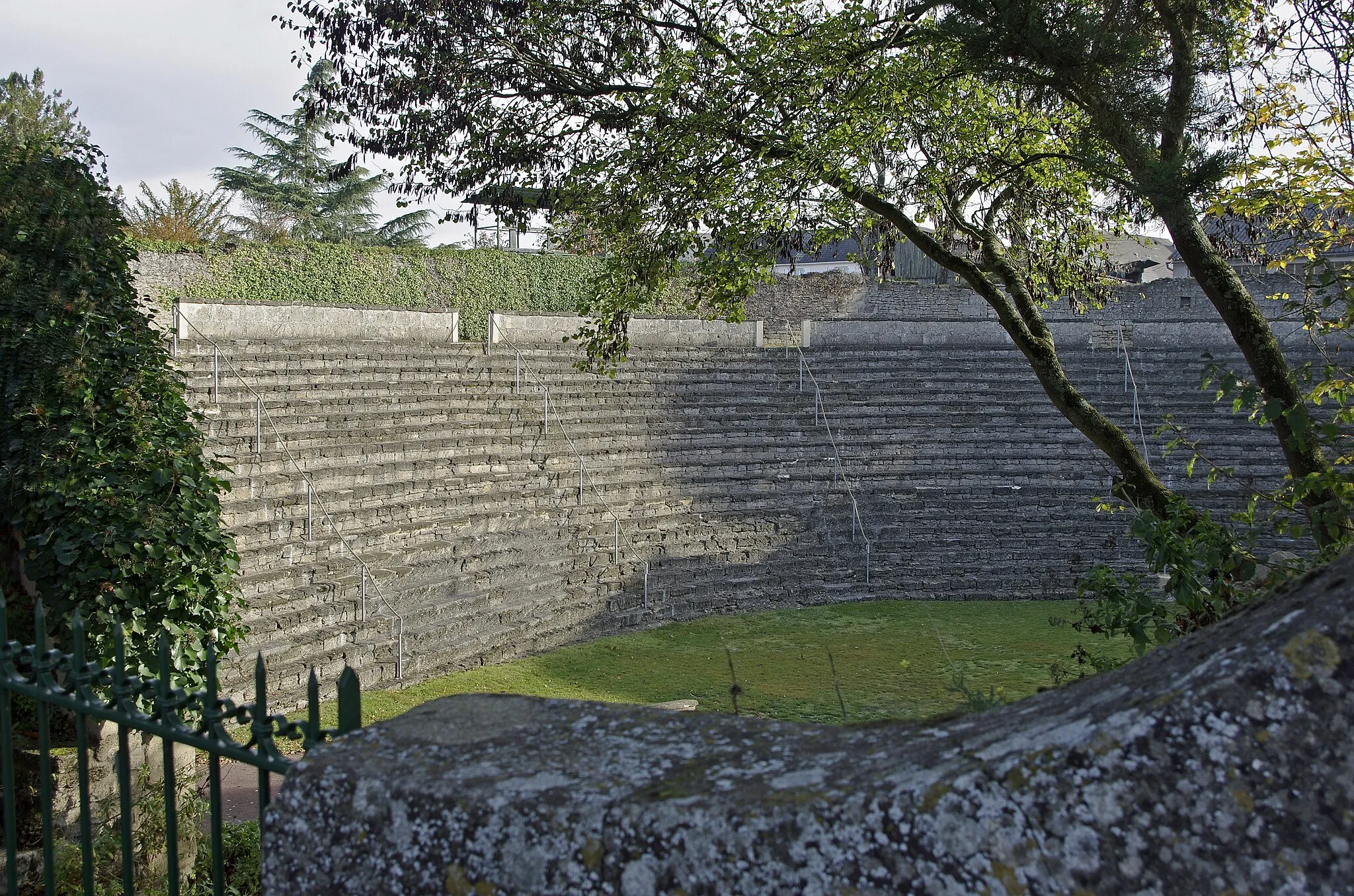 Photo showing: Doué-la-Fontaine (Maine-et-Loire)
Les Arènes.
Elles ne sont pas gallo-romaines. Elles proviennent de la transformation d'une carrière à ciel ouvert en amphithéâtre. Elles sont probablement pas antéreures au XVe siècle.
Au XVIe siècle, Rabelais mentionne à plusieurs reprises dans sont Pantagruel, les "diableries"* qui se déroulent dans le Parc des jeux de Doué*.
Le spectacle dans les arênes semble avoir cessé en 1750.
Durant les guerres de Vendée, le site servit de prison. Doué fut le théâtre de batailles: prise par les Vendéens le 7 juin 1793, reprise par les Bleus le 5 août suivant. Près de 200 Vendéens furent enfermés dans les arênes à la fin de l’année 1793.
Les arènes servirent de cantonnement militaire pendant la guerre de 1870, puis d'auberge, de dépendances agricoles, de dépotoir.
En 1910, une société fut créée et les Arènes redevinrent un lieu de spectacle.
Pendant la seconde guerre mondiale, les Allemands entreposèrent leurs munitions dans les caves.
Le site sera racheté par la municipalité en 1952, et aujourd'hui, il accueille des représentations et manifestations, tel que le Festival d'Anjou, en juin.

Les "diableries" sont des parodies des Mystères de la Passion. A partir de la fin du XIIIe siècle, le thème principal des "Mystères" (genre de théâtre médiéval), est celui de la Passion de Jésus-Christ. A la fin du XVe siècle, le tourangeau Jean Michel présente une grande Passion qui compte près de 34 000 vers et dont la représentation s'étalait sur une dizaine de jours. Jean Michel y multiplie les épisodes truculents, avec des scènes de bouffonnerie pure. Il y décrit la vie mondaine de Madeleine et les amours de Judas. Cette Passion de Jean Michel annonce les farces scatologiques et obscènes des bateleurs, ce qui amènera le Parlement de Paris à interdire les Mystères en novembre 1548. "De cestuy monde rien ne prestant ne sera qu'une chienerie, que une brigue plus anomale que celle du recteur de Paris , qu'une diablerie plus confuse que celle des jeux de Doué,... (Livre III. Chap. III.) Meffieurs les diables, vous jouërez bien, je vous affie. O que vous jouërez bien. Je defpite la diablerie de Saulmur, de Doüé, de Mommorillon , de Langes , de fainct Efpain, d'Angiers : voire , par Dieu, de Poictiers avecques leur parloüoire, en cas qu'ils puiffent eftre à vous parragonnez. (Livre IV. Chap. XIII.)
Nos petites caroles et puériles esbatemens achevez, houstans nos faulx visaiges, appareumes plus hideux et villains que les diableteaux de la Passion de Doué, tant avions les faces guastées aux lieux touchez par les ditz feuilletz. (Livre IV. Chap. LII.)

www.ville-douelafontaine.fr/user/documents/fichiers/vivre...