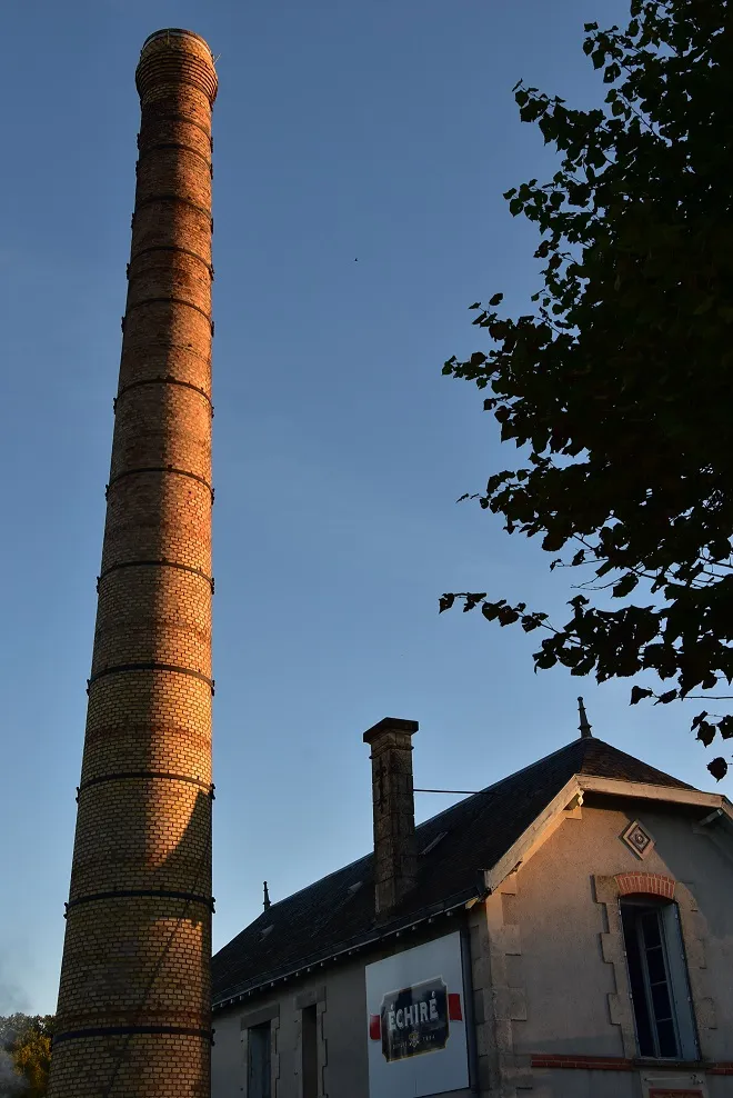 Photo showing: Coopérative laitière de la Sèvre à Echiré (79) et sa cheminée de briques caractéristique, face à l'église communale.
Le site voit le jour en 1893 et prend son nom actuel en 2004, suite à sa fusion avec Sèvre & Belle, implantée à Celles.

Elle fait travailler 120 professionnels locaux situés dans un rayon de 50 km, presque équitablement répartis entre lait de vache et chèvre. Les produits, beurre, crème fraiche et fromages en tête, sont diffusés à l'international à travers le circuit des épiceries de luxe.