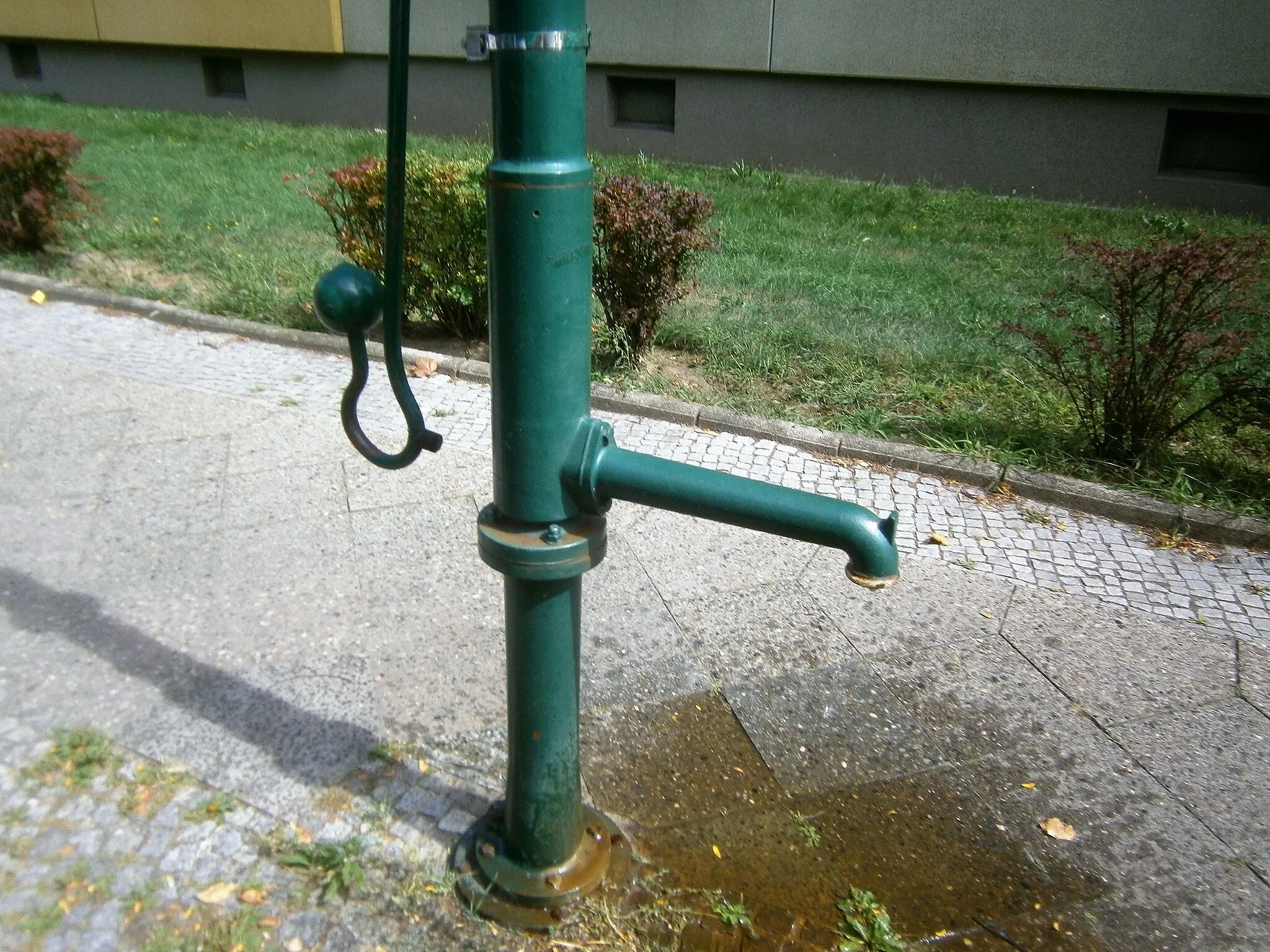 Photo showing: Der Standort des Brunnens zwischen Haakezeile und Pfefferweg ist mit Hausnummer 8 (Wohnzeile 4–8) in der Liste benannt. Ein FSH-Brunnen steht 2008 am östlichen Gehweg, allerdings vor dem viergeschossigen Wohnhaus 16 (Wohnzeile 16–20). Der (vermeintliche) Abstand beträgt dabei 60 Meter, wobei zwischen dem Haus 8 und 16 die Zufahrt zum querstehenden Block 10–14 liegt, sowie die Müll-/ Recyclingcontainer und eine Garagenreihe.[1] Die Wohnhäuser im Karree wurden in den 1960er Jahren erbaut, der Brunnenstandort könnte wohl zu diesem Zeitpunkt festgelegt sein. Der Brunnen hat für den Wasseraustritt eine lange Hülse am Schaft. Der Abfluss erfolgt unmittelbar auf den Gehsteig. Um den Brunnenfuß, der am Flansch des Sockelrohrs verschraubt ist, liegt in einer Kleinpflasterung.