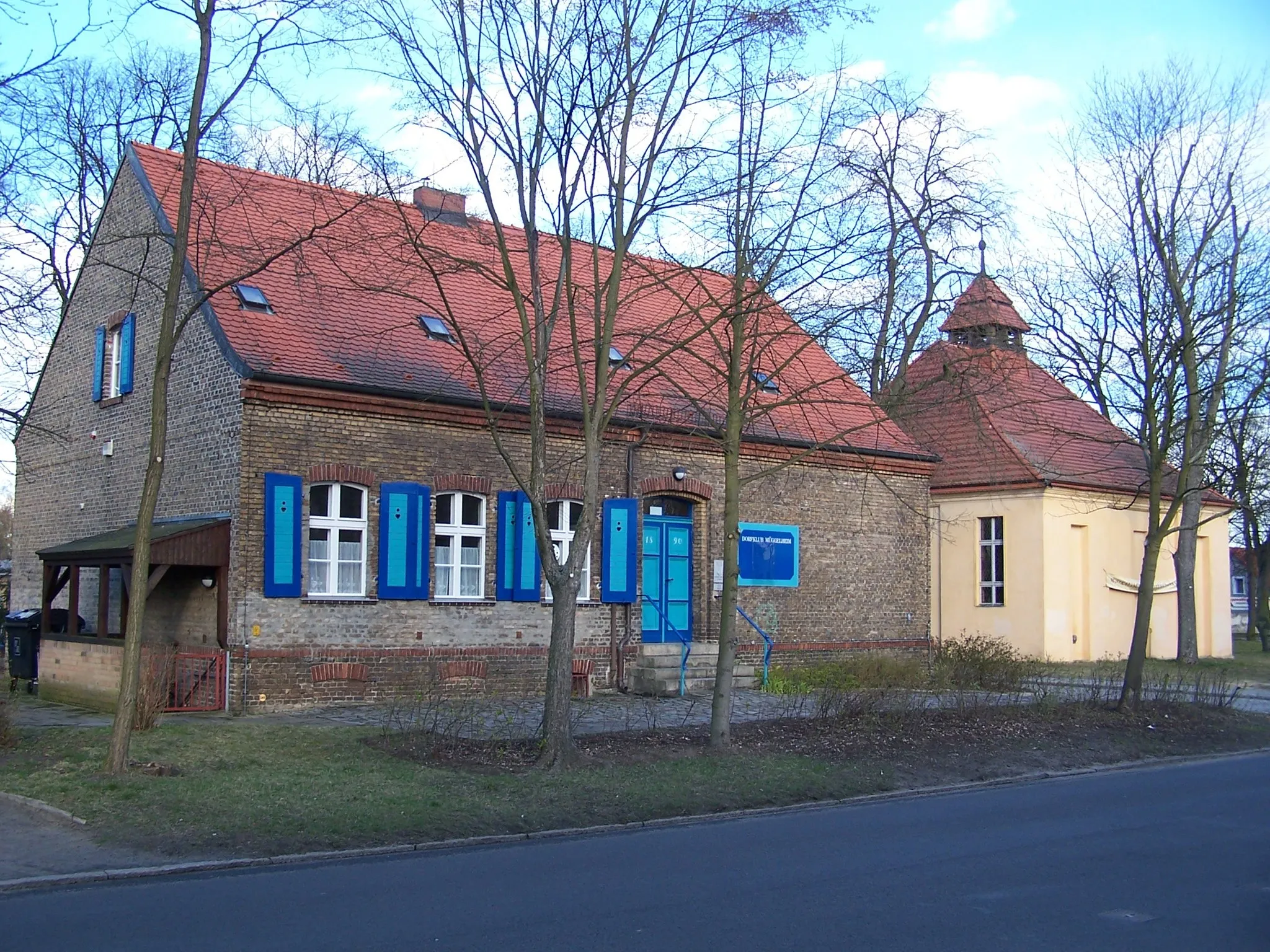 Photo showing: The building of the former school of Berlin-Müggelheim (which serves as the “Dorfclub“ (clubhouse) today) and the church in the background.