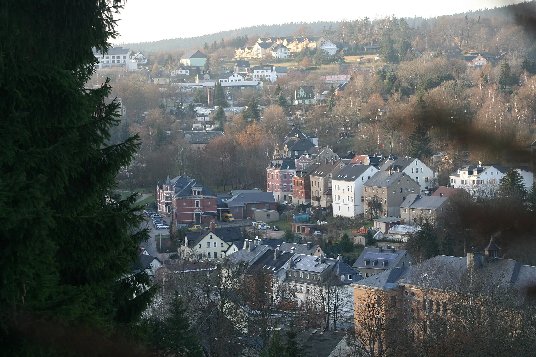 Photo showing: Schönheide im Erzgebirge: Vom Denkmal am Knock geht der Blick über Mitteldorf und Ortsteil Fuchswinkel zum Fuchsstein. Rechts das imposante Gebäude der Geschwister-Scholl-Schule, die von 1896 bis 1998 gebaut wurde. Die roten Ziegelbauten links sind vorn die Post von 1891 und dahinter das Hotel zur Post etwa aus der gleichen Zeit.  Mehrfamilienhäuser aus der Gründerzeit und später zeigen die stadtähnliche Bebauung aus der Zeit der Verstädterung. Neuere Bauten aus den 1990er und 2000er Jahren finden sich im oberen Bildbereich am Fuchsstein. Fotografiert vom Berg Knock aus.