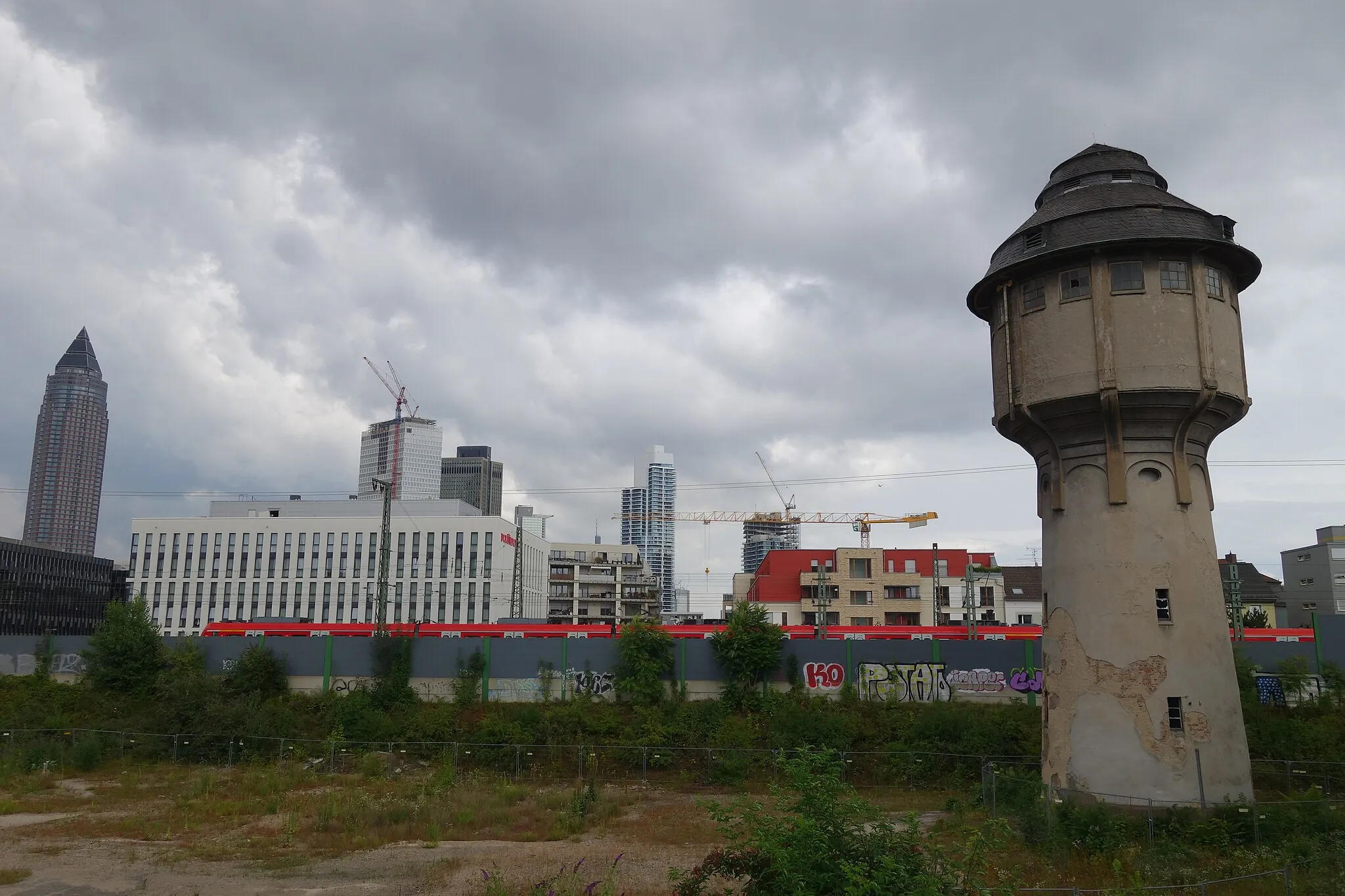 Photo showing: Alter Wasserturm des einstigen Hauptgüterbahnhofs Frankfurt, von Westen gesehen. Hinter der Schallschutzmauer fährt ein S-Bahn-Triebzug der DB-Baureihe 423 auf der Main-Weser-Bahn zwischen Frankfurt-Messe und Galluswarte. Im Hintergrund von links die Hochhäuser Messeturm, One (im Bau), Tower 185, "Kronenhochhaus" (bzw. Westendstraße 1), Grand Tower und Eden (im Bau