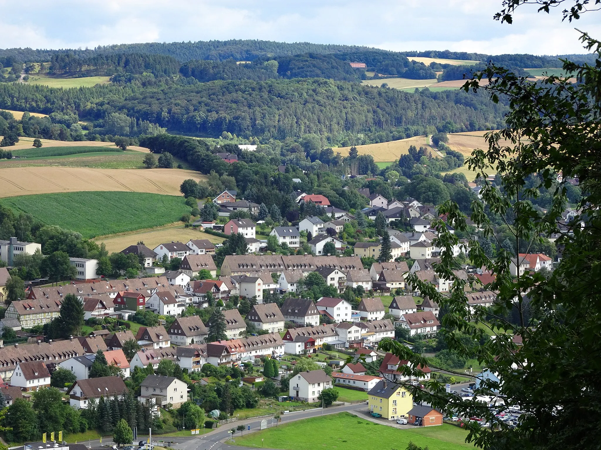 Photo showing: Sontra im nordhessischen Werra-Meißner-Kreis.
Die frühere (Bergbau-)Siedlung II.
Oben die "Schulstraße" darunter die parallel verlaufende "Knappenstraße".
Ganz unten die ehemalige Bundesstraße 27 und nach dem Bau der Umgehung, die heutige Innerortsstraße "Göttinger Straße"
" Gesamtanlagen Barbaraplatz und Glückaufplatz
In der Mitte der dreißiger Jahre des 20. Jahrhunderts sollte der Kupferschieferbergbau im Richelsdorfer Gebirge erneut aufgenommen werden, nachdem die Arbeiten 1870 eingestellt worden waren. Zu diesem Zweck mussten Wohnmöglichkeiten für die zuziehenden Bergleute geschaffen werden, da die Bevölkerungszahl Sontras von 2400 Einwohnern im Jahr 1937 bis auf 5000 Einwohner im Jahr 1939 ansteigen sollte.
Um den ersten Ansturm der angeworbenen Bergleute aus Schlesien und dem Saarland aufzufangen, wurden zwei Neubausiedlungen geplant. Die Konzeption, die im Dezember 1936 erstellt wurde, sah zwei getrennte Siedlungen am Stadtrand vor. Die Bebauung im Bereich des Barbaraplatzes sollte über 360, diejenige des Glückaufplatzes über 216 Wohneinheiten verfügen. Die Planungen gingen von einer Siedlungsstruktur aus, die sich um eine Mittelpunktfunktion einnehmende Platzeinheit mit einem zentralen, gemeinnützigen Gebäude konzentrierte.
Die Straßenzüge, deren Zeilenbebauung von unterschiedlichen Haustypen geprägt wird, sollten sich dem Verlauf des umgebenden Geländes einfügen. Fachwerkarchitektur wechselt mit massiven Gebäuden, die jeweils in lockerer Gruppierung einander zugeordnet sind.

Nach dem zweiten Weltkrieg kamen die Häuser in die Hände der halbstaatlichen Kurhessen Wohnungsbau GmbH, die privater Kundschaft Kaufmöglichkeiten eröffnete. Diese Privatisierung bedingte Eingriffe in die Bausubstanz, die die Einheitlichkeit des Ensembles jedoch kaum beeinträchtigten."