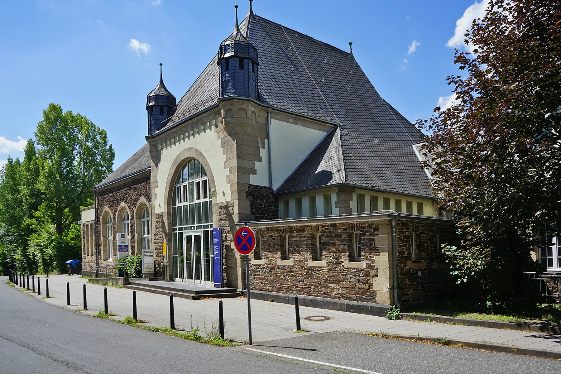 Photo showing: Bullay (Mosel) - Bahnhof, Empfangsgebäude (1904) Straßenseite
Bullay, Rheinland-Pfalz, Juli 2020
Der Bahnhof Bullay entstand mit dem ab 1875 begonnenen Bau der Moselstrecke von Koblenz nach Trier, die am 15. Mai 1879 eröffnet wurde. Das erste Stationsgebäude war ein einfacher Holzfachwerkbau. 
Das heutige Empfangsgebäude wurde 1904 durch ein größeres Gebäude im Heimatstil mit Bahnhofsrestaurant und Paketabfertigung ersetzt (Entwurf: Alexander Rüdell (1852-1920)).
Um 1910 diente die Bahnhofshalle mit Jugendstilfliesen aus Ehrang dem Empfang und Service wie Fahrkartenschalter und Gepäckausgabe. Die tonnengewölbte Decke des Bahnhofsrestaurants war mit Schablonenmalereien im Jugendstil verziert, und das verwendete Geschirr trug die Aufschrift "Staatsbahnhof Bullay".
Nachdem das Transportaufkommen und die Fahrgastzahlen nach dem Zweiten Weltkrieg zunehmend rückläufig waren, wurde Bullay 1995 neben drei weiteren Bahnhöfen für das Projekt des „Umweltbahnhofs des Landes Rheinland-Pfalz und der Deutschen Bundesbahn“ aus 38 rheinland-pfälzischen Bahnhöfen ausgewählt.
Dies ermöglichte eine umfassende Neugestaltung mit dem Ziel, die Umweltverträglichkeit des Verkehrsmittels Bahn herauszustellen. Im Rahmen der umfangreichen Bauarbeiten ab 1999 wurde auch das historische Empfangsgebäude größtenteils im ursprünglichen Stil renoviert und alle sonstigen Bahnhofsbauten abgerissen.
Der Bahnhof wurde zur Etappe des Moselradweges mit der Möglichkeit, Fahrräder unterzustellen, zu duschen und die Toiletten zu benutzen. Die Neueröffnung erfolgte zu Pfingsten 2003.
Quellen:
Wikipedia (de.wikipedia.org/wiki/Bahnhof_Bullay_(DB))

KuLaDig, Kultur.Landschaft.Digital. (www.kuladig.de/Objektansicht/O-139649-20150917-2)