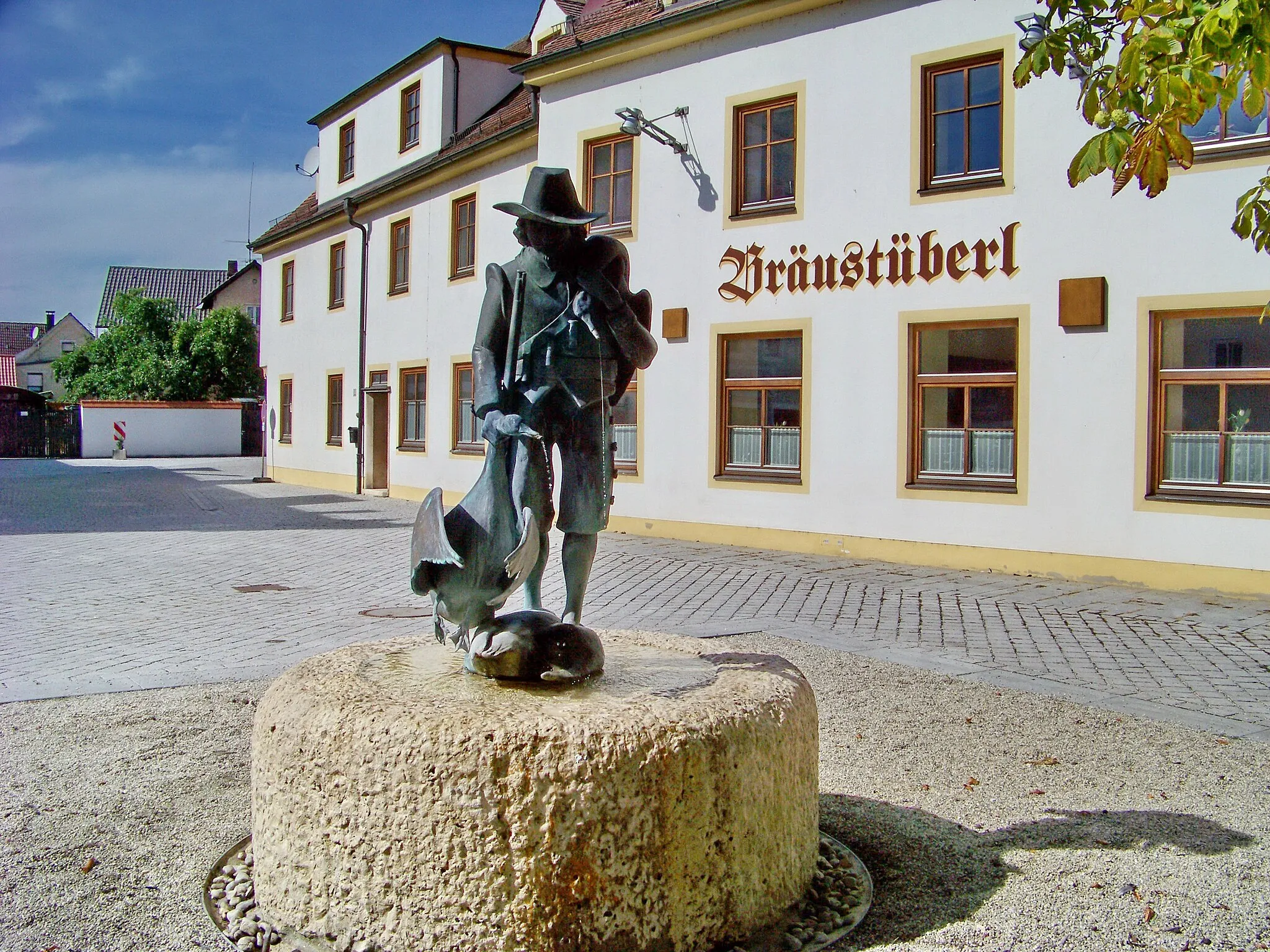 Photo showing: Im Markt Schierling steht der sogenannte Gänshängerbrunnen am Rathausplatz. Die Bronzeskulptur geht auf ein geschichtliches Ereignis im 30-jährigen Krieg zurück. Es herrschte große Not. Damit seine Gänse nicht in die Hände der plündernden Schweden fallen, schlachtete der Schlossherr sie und hängte sie an einen Haken unter einer Brücke. Ein schwedischer Musketier fand die Gänse und es gab einen festlichen Braten. Aus den Schnäbeln der Gänse fließt Wasser. Die Figur zeigt den schwedischen Soldaten. Die Skulptur wurde vom Bildhauer Klaus Vrieslander im Jahr 1980 geschaffen.