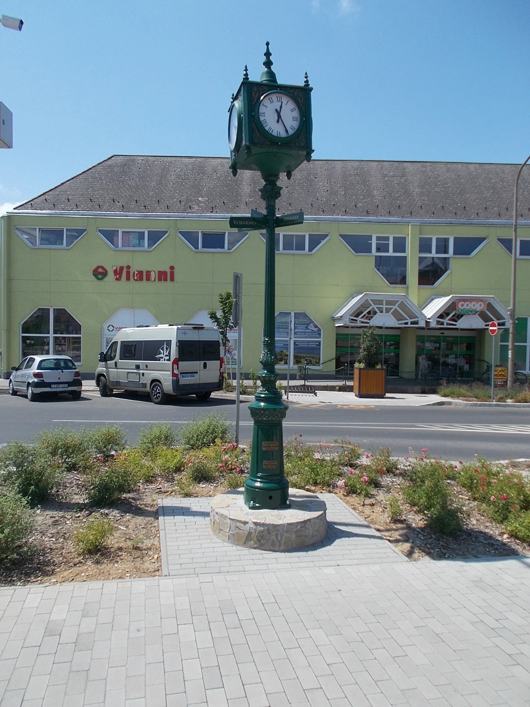 Photo showing: Street clock made by Szabados and Associate (from Budapest). Twin towns direction and name signs on the column. At back a Vitamin/probably Nature food shop - Flórián Square, Sümeg, Veszprém County, Hungary.