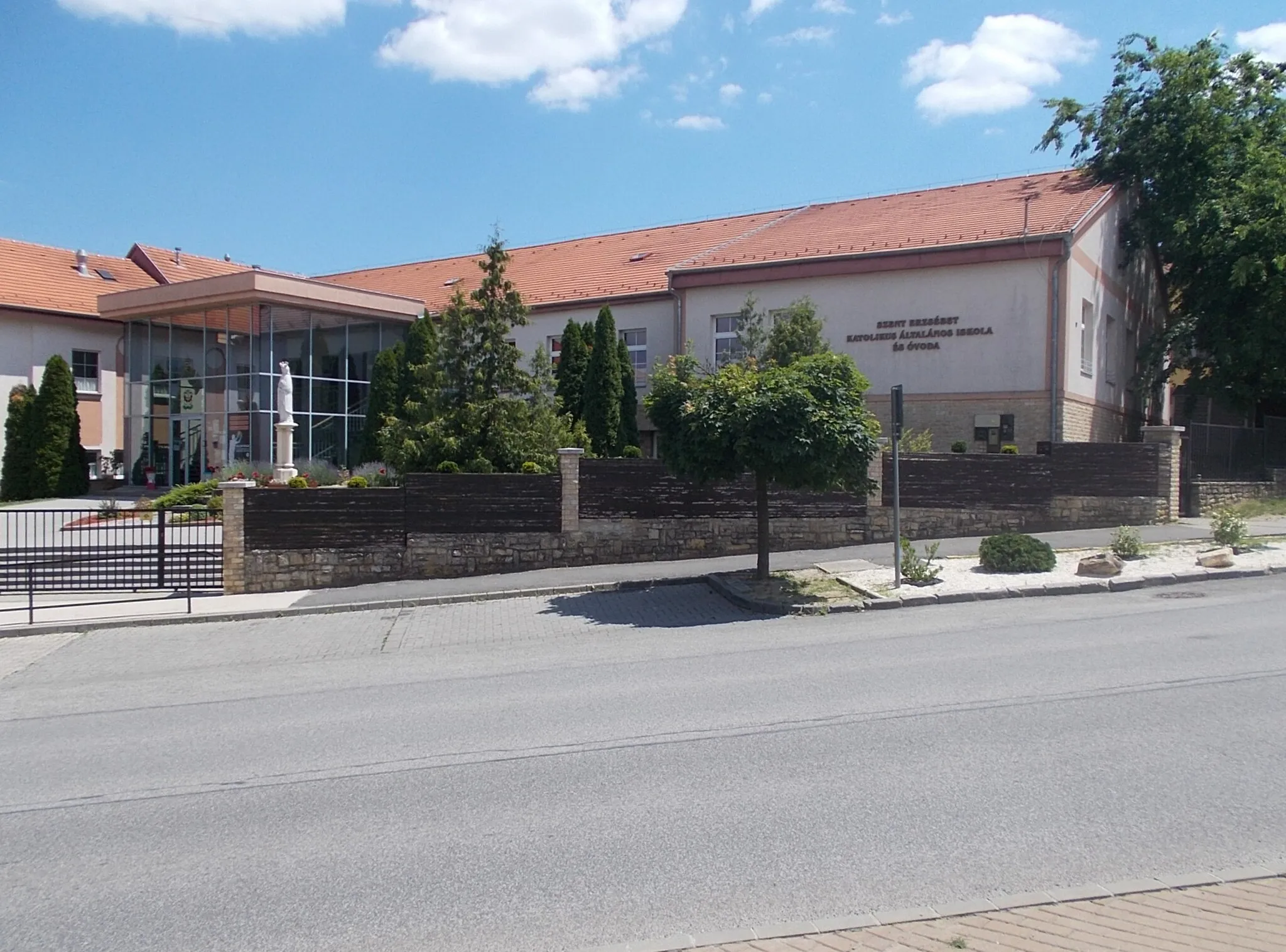 Photo showing: St. Elizabeth's Catholic Primary and Kindergarten. Inside in the aula is a St. Elizabeth relief by Károly Fésűs (2008 gypsum?? works, size 5m x 2.5m) and outside in the street side garden/courtyard is a St. Elizabeth by Károly Fésűs (2003 statue, which is about 1.30m high, stands on a 1.70m high pedestal, decorated with three columns) - 7 Kossuth Square, City centre, Pécel, Pest County, Hungary.