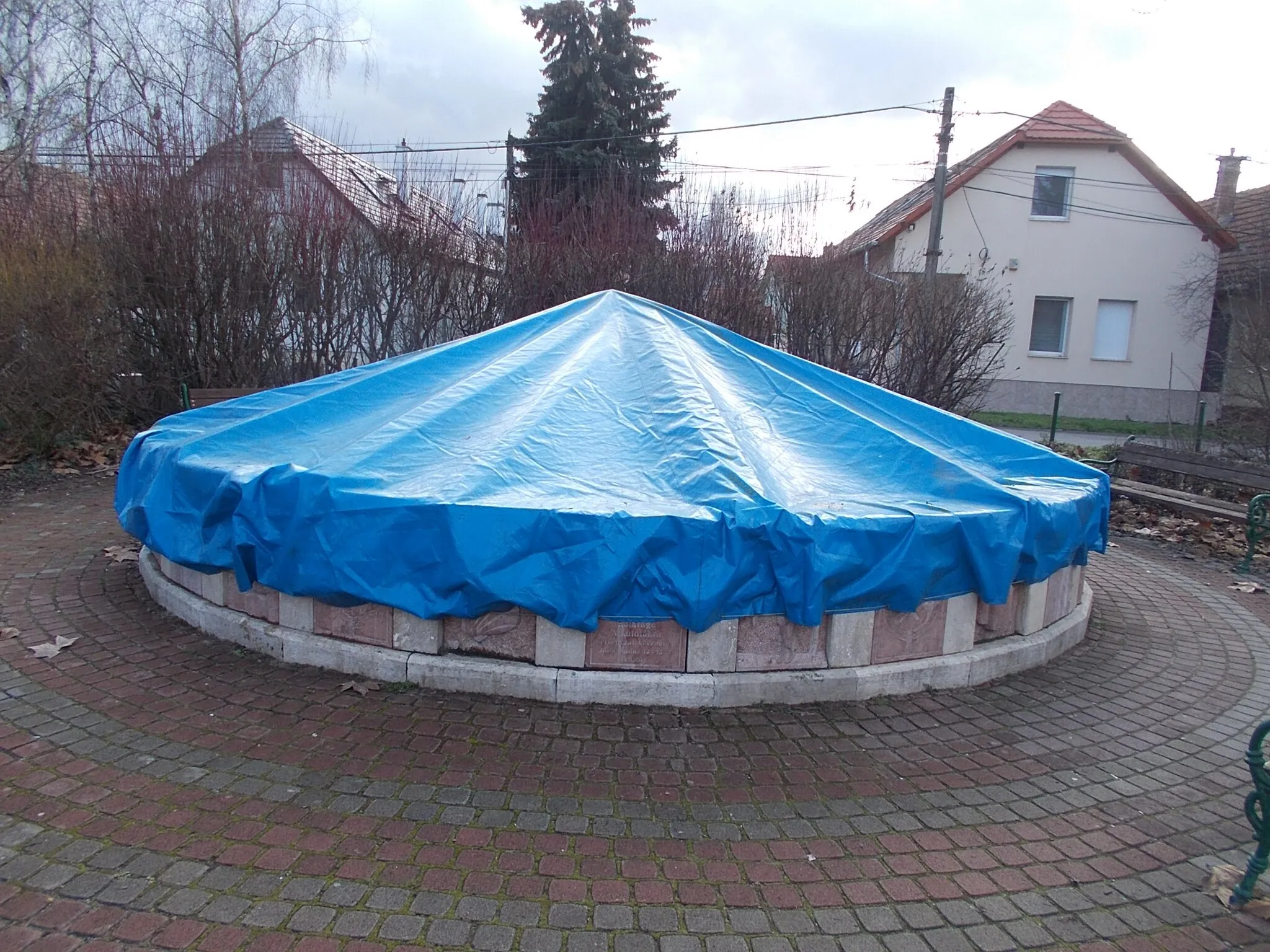 Photo showing: Fountain wrapped/covered for cold/snow protection. Built in 2008. Its bassin/pool decorated with 50x40x12cm size reliefs The reliefs made by Participants of the Central European Stonemasonry Creative Camp. Creators and their creations: Andrea Nagy from Majosháza (mermaid), Roland Ugodi from Siklós (aquarius), Gergő Flock from Sződ (dolphin), Antal Plank Jr. from Érd (dried-up riverbed), Róbert Arnhoffer from Márkó (seahorse) and Gergely Novák from Alsóörs (pearl mussel), Verguere Paul Joseph from France (devil of the sea) - Fő Street Park middle, Üröm, Pest County, Hungary.