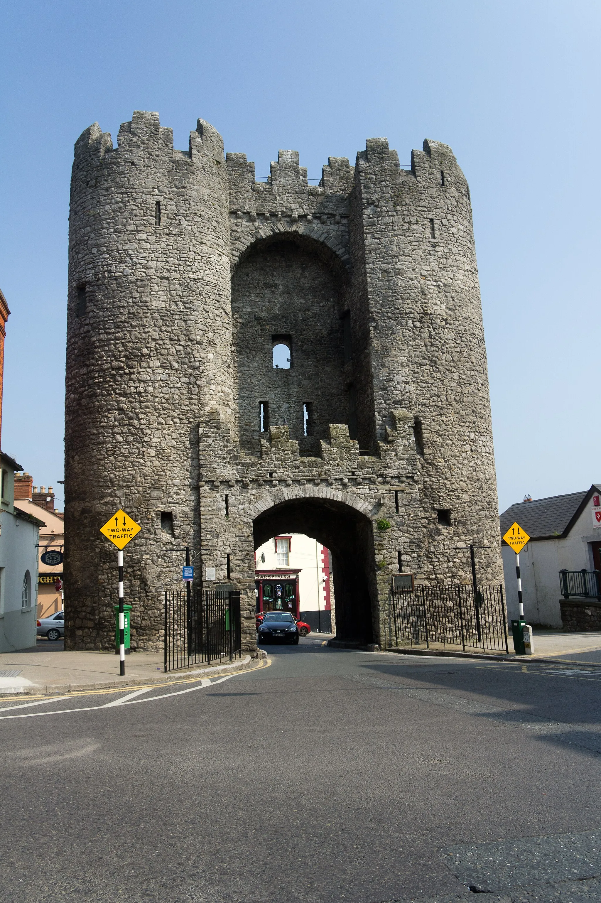 Photo showing: Laurence's Gate is a barbican which was built in the 13th century as part of the walled fortifications of the medieval town of Drogheda, County Louth, in Ireland. The original names for Laurence St and Laurence's Gate were East St and East Gate, respectively. In the 14th century, the street and Gate were renamed because they led to the hospital of St. Laurence, which stood close to the Cord church.
It consists of two towers, each with four floors, joined by a bridge at the top and an entrance arch at street level. Entry is gained up a flight of stairs in the south tower. There is a slot underneath the arch from where a portcullis could be raised and lowered.
Historians have wondered why such an enormous barbican was built here in the east of the town, when the main artery through the town has always been north/south. For example, a similar barbican in Canterbury is less than half the height of Laurence's Gate. However, from the top of the Gate, the estuary of the Boyne and the four mile stretch of river from there to Drogheda can be clearly observed. This is the only point in the town with a clear view of any potential sea invasion. This is proposed as a reason why Lawrence's Gate was built to such a height.

A portion of the town wall remains to the south of Laurence's Gate. North of Laurence's Gate, the wall ran up Palace St/King St where the footpath is today. The depth of the basements of the houses and school suggest the presence of a steep trench outside the wall. Over the centuries, as the walls and gates fell into disrepair, the rubble stones were reused in later buildings. For example, the house and walls at the corner of Laurence and Palace St and stone walls in Constitution Hill. Old pictures show that a toll booth and gate house remained until the early 19th century. The shop beside Laurence's Gate was a bicycle shop 100 years ago. The green letter box dates from a time when there was a post office there.