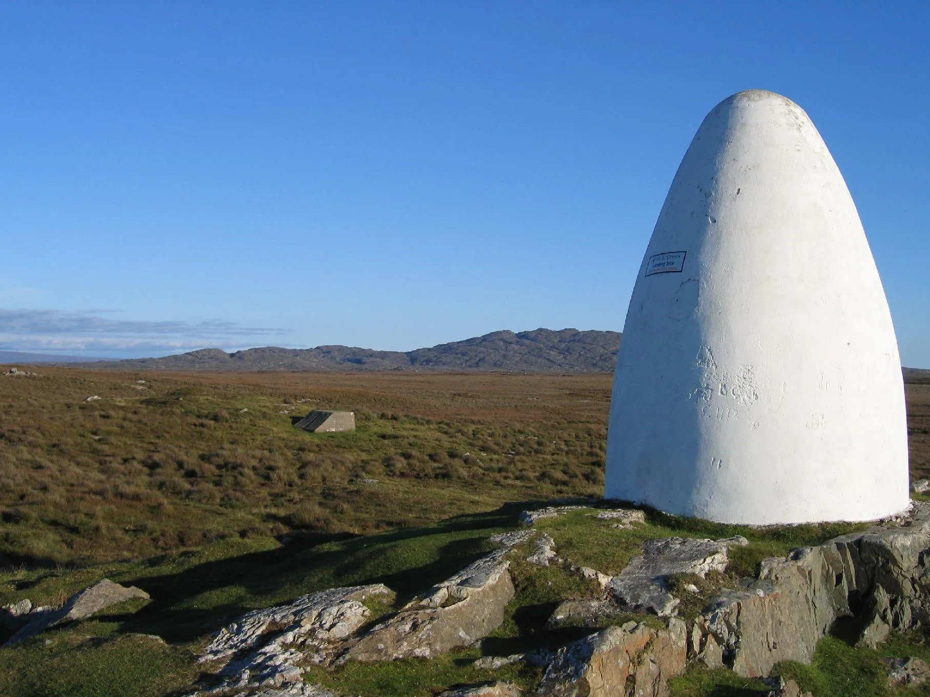 Photo showing: The landing site of the pioneering transatlantic flight of Alcock and Brown, and adjacent memorial cairn.  The first non-stop transatlantic flight crash-landed four kilometres south of Clifden, County Galway, Ireland on June 15 1919. There is a memorial cairn around four metres in height on the site of Marconi's first transatlantic wireless station, five hundred metres from the crash site. The inscription on the cairn reads "Alcock and Brown, landing site, 500 meters" with an arrow pointing towards the bog in the centre of the picture.