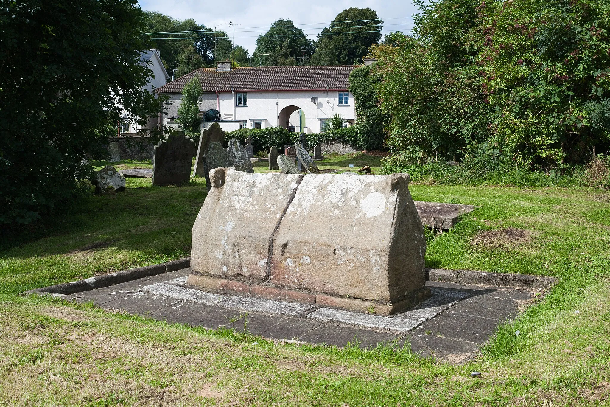 Photo showing: Shrine in form of a wooden church close to the round tower, considered by Michael Moore as possibly the oldest surviving structure of St Tigearnach’s Monastery. W. F. Wakeman describes it as follows: “Situated upon a line between the round tower and the abbey, and directly facing the doorway of the former structure, occurs a monument, in some respects unlike anything which has elsewhere been noticed in Ireland. It is formed of a single block of hard red sandstone, 5 ft. 10 in. in length by 3 ft. in height, which has been fashioned into the form of an early Irish church. The interior is artificially hollowed, so that the work forms a shrine in form exactly like that of St. Ethelreda, preserved in Ely Cathedral. Its position is east and west, like that of a church. Upon what I may style the eastern gable is carved the upper portion of a human figure, the head of which is covered by a triangular head covering, probably a mitre. Upon the southern side, just below the eve, are two heads of animals, graven bracket-fashion, and very similar in style to some of the grotesque carvings remaining within the round tower of Ardmore. This side, also, bears an unintelligible sculpture [..]. The northern and western sides are quite plain. It may be observed that the tops of the gables are finished much in the same manner as those of the representations of early churches which we often see terminating our richer crosses.” The shrine was used for burials in post-medieval times where, according to local tradition, the shrine was used as a family grave by the McMahons or the McDonnells. This was described as follows: “It [the shrine] is not so small. It gets wide towards the floor. If the copestones were off, two or three men could move about in it. Besides, the mode of interment was this: the corpse was taken out of the coffin, and left on ‘stretchers,’ the empty coffin left beside it; lime was then applied. When the next corpse came, the former empty coffin was taken out and broken beside the tomb.” See the record of the Archaeological Survey of Ireland and Wakeman, W. F. “On the Ecclesiastical Antiquities of Cluain-Eois, Now Clones, County of Monaghan.” The Journal of the Royal Historical and Archaeological Association of Ireland, vol. 3, no. 21, 1875, pp. 327–340. Fourth Series, www.jstor.org/stable/25506666.