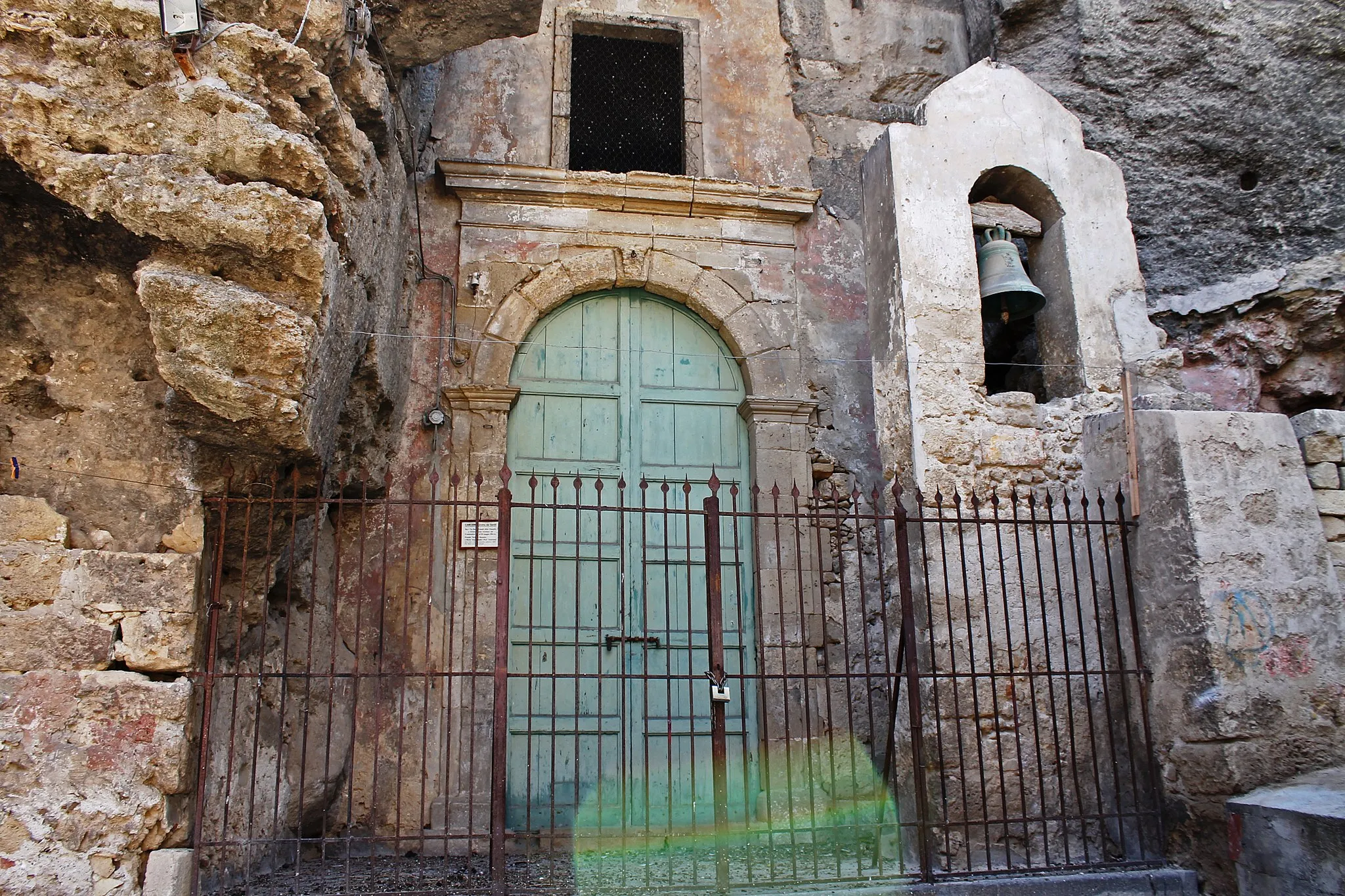 Photo showing: La Chiesa del Sacro Carcere dei Tre Santi è un luogo molto importante per il culto legato ai "Tre Martiri di Lentini". Essa si trova presso la Via Paradiso( di preciso in "Vicolo tre santi").
Questa chiesa è nota come: "Grotta dei Santi", "Chiesa di San Giuliano Spadaliere Patrono dei Viandanti" e con i nomi siciliani di "Rutta , Rattatullu", cioè "Grotta di Tertullo" (colui che fece uccidere i tre Santi) oppure di "A Rutta 'e Santi" (La Grotta dei Santi). In verità essa è una piccola caverna, utilizzata in età greco - romana come carcere. In questa grotta vennero incarcerati e torturati i santi Alfio, Filadelfo e Cirino, ma qui vi furono anche molte apparizioni di "Cristo" ai tre fratelli e anche numerose guarigioni.

L'ingresso della chiesa di forma arcuata è delimitato da una cancellata in ferro battuto accanto alla quale vi è una piccola torre campanaria. All'interno vi è la grotta (ora adibita a chiesa) dove nei giorni della festa patronale vi è ricostruita la "Carcerazione di Alfio, Filadelfo e Cirino" con pregevoli statue. Sull'Altare Maggiore vi sono poste le statue che raffigurano i tre fratelli, mentre a sinistra vi è situata la statua raffigurante santa Tecla (Compatrona di Carlentini), colei che aiutò i tre fratelli durante la loro prigionia, come ringraziamento per il miracolo compiuto su di lei ( Tecla non riusciva più a camminare, ma bastò un tocco dei tre fratelli per guarirla). Bisogna dire che questo è un esempio ben conservato di "Chiesa rupestre paleocristiana", che conserva anche alcuni interessanti ipogei che videro la sepoltura di molti martiri.