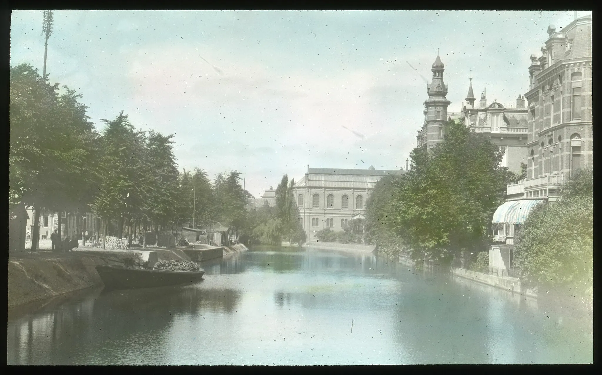 Photo showing: View looking down a canal in Amsterdam. Several boats sit moored at the side of the canal opposite a tall spire topped building. The Arrases visited Amsterdam during their 1913 tour of Europe. Note on slide reads "Amsterdam canal." Edmund F. Arras (7/7/1875-10/19/1951), a prominent Columbus businessman and entrepreneur, founded one of Columbus, Ohio’s first property rental agencies in 1892. Trained as a lawyer, he graduated from OSU law school in 1896 and went on to hold positions in numerous civic groups around the city. He was particularly active within Kiwanis International and was involved with several local religious organizations. In 1913, he and his wife Elizabeth traveled to the World Sunday School Association Convention in Zurich, Switzerland. They continued on to travel extensively throughout Europe documenting their journey through photographs. Due to the timing of their trip, these photos comprise a valuable collection of images of European cities later devastated by war. The Arras family’s lantern slides and negatives include images from their 1913 travels through US east coast cities, Atlantic islands of the Azores, Madeira, Gibraltar and the countries of Algeria, Italy, Switzerland, Germany, the Netherlands, Belgium, France, England, and Ireland. Also included are images from their 1920 train journey from Chicago to the western US for the Kiwanis Convention in Portland. These photographs include images of Chicago, Denver and western National Parks. Negative image also in collection HA3_B10_0545.
