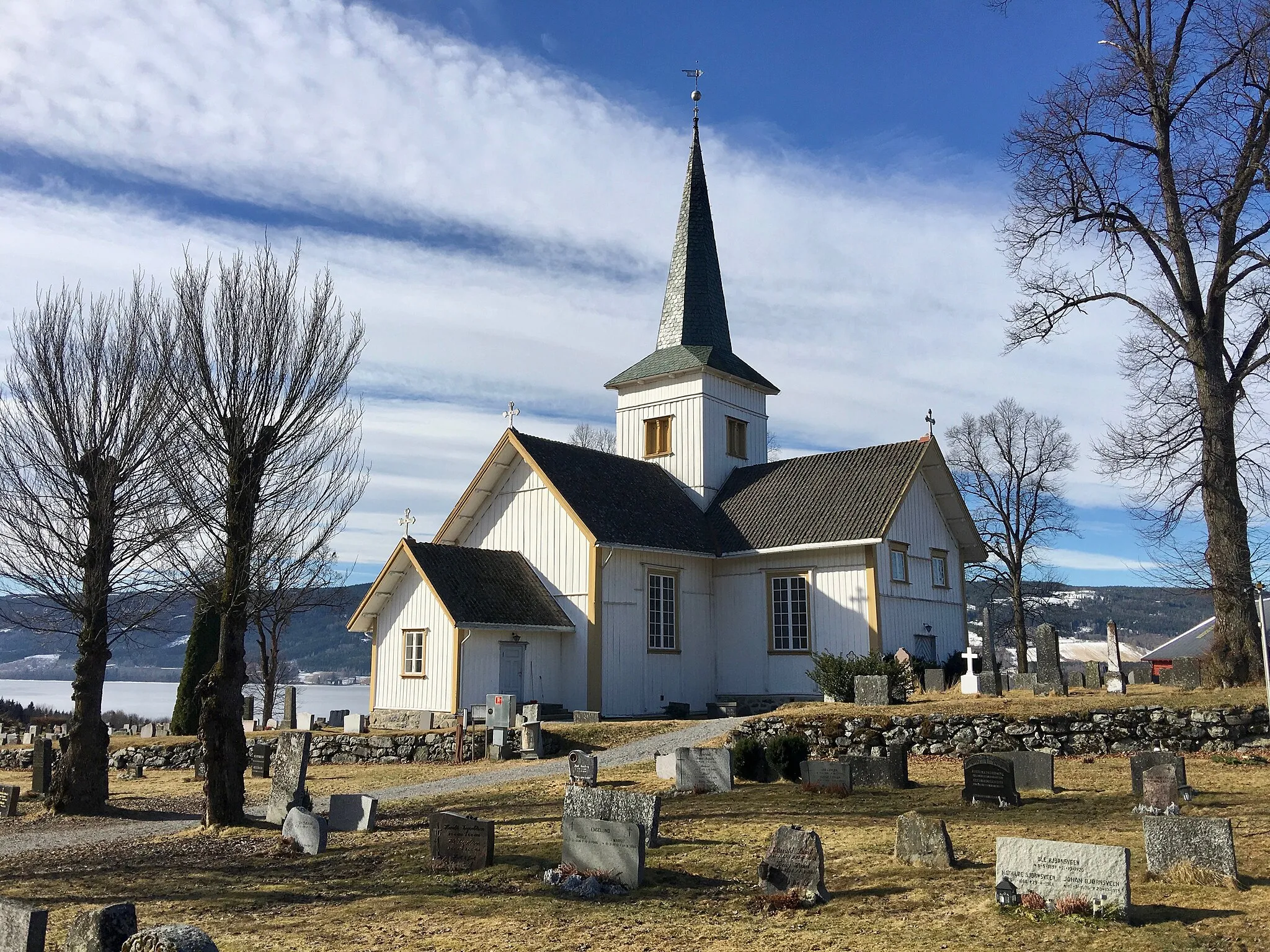 Photo showing: Hov Church (Hov kirke, built 1781) in the village of Hov in Søndre Land Municipality, Oppland County, Norway. The photo shows the cemetery /graveyard (kirkegården) surrounding the wooden church with naked trees in spring, a view towards the randsfjorden lake, etc. Photo taken on March 27th, 2017.