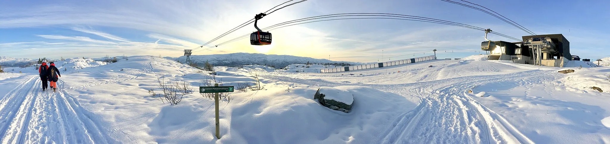 Photo showing: The top station of the Voss Gondol, an aerial passenger lift (gondolheis, taubane) opened in 2019 in the small town of Vossevangen in Voss municipality in the western part of Norway. The cableway goes from the Voss Railway Station to the summit of Hangur/Hanguren mountain 820 metres above sea level, north of the town, with the Hangurstoppen Restaurant, a ski resort, hiking aera, paragliding site, etc. The gondola lift has nine cabins.
Distorted panoramic photo taken on November 20th, 2019. The afternoon sunset colours the snowy landscape yellow and blue due to the low sun and long shadows. Also seen are two paraglider pilots going to the Bavallen flying site, sign for the Panoramaløya hiking route, etc.