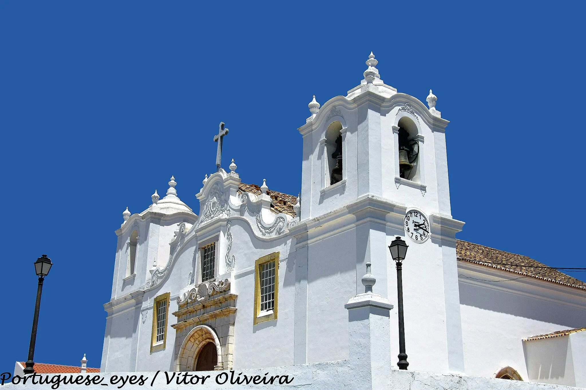 Photo showing: De remota fundação, a Igreja Matriz da bonita povoação de Estômbar, que integra o concelho da Lagoa, foi remodelada no reinado de D. Manuel I. No entanto, o século XVIII havia de a reformar quase totalmente, conservando este templo ainda alguns sinais de épocas anteriores. A Matriz de Estômbar é dedicada ao apóstolo S. Tiago e foi decretada em 1984 Monumento Nacional. Situada no Largo da Igreja desta povoação algarvia, a Matriz de S. Tiago de Estômbar ergue-se no topo de um morro, ao qual se acede por escadório de vários patamares.
Antecedida por um adro, a imponente frontaria revela a reforma da segunda metade do século XVIII. Na fachada rasga-se um belo portal quinhentista da época manuelina, em arco de volta perfeita, enquadrado por estrutura de pedra idêntica (grés escuro local); as suas colunas apresentam decoração naturalista e curiosos e invulgares motivos antropomórficos - músicos com os seus instrumentos e figuras femininas, talhados com alguma rudeza, mas imbuídos de uma rica sensibilidade popular.
A estrutura do portal é encimada por movimentadas aletas, sobre as quais se rasga uma janela retangular, ladeada por motivos barrocos curvilíneos. A porta é flanqueada por duas janelas retangulares gradeadas. A empena desenha linhas ondeadas, encimada por frontão curvo, pináculos e cruz latina, no tímpano do qual se inscreve um medalhão barroco ornamentado. A fachada é flanqueada por duas torres sineiras quadrangulares, marcadas por cunhais salientes e divididas em dois pisos. O superior é rasgado por ventanas, que apresentam cobertura curva marcada por várias urnas sobre pedestais.O interior é formado por três naves, repartidas em quatro tramos por arcos de volta perfeita, forrados por painéis de azulejos setecentistas. As paredes laterais das naves apresentam idêntico revestimento cerâmico.
Sobre o arco triunfal e circundando um óculo, expõe-se um painel cerâmico alusivo à Ascenção. A capela-mor é totalmente forrada com azulejos setecentistas (1719), figurando episódios hagiográficos e graciosos anjos brincando. O retábulo-mor é uma composição do barroco tardio, decorado com três faustosos medalhões e ostentando num dístico a data de 1760. In Infopédia [Em linha]. Porto: Porto Editora, 2003-2011. [Consult. 2011-10-29].
Disponível na www: .

See where this picture was taken. [?]