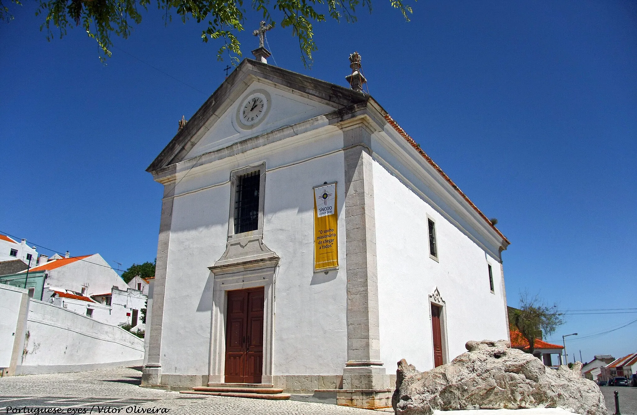 Photo showing: Fundada no século XIV, a igreja paroquial de Vialonga foi objecto de várias campanhas de obras (principalmente no século XVI) que lhe conferiram o seu aspecto actual, um volume compacto, onde impera a ausência de elementos decorativos. Apenas a fachada é animada por um portal rectilíneo e pela janela do coro, sendo rematada por frontão triangular com relógio no tímpano. Do lado da torre sineira, num plano mais recuado, encontram-se as dependências anexas.
Esta austeridade arquitectónica contrasta fortemente com a decoração do interior, que tira partido da conjugação da talha, da pintura e do azulejo, num repositório muito caraterístico da primeira metade do século XVIII. O espaço, de nave única coberto por abóbada de berço, articula-se com a capela-mor, profunda, através de um arco triunfal de volta perfeita, flanqueado por dois altares de talha dourada seiscentista. Para além do púlpito de mármore com balaústres, no corpo do templo abrem-se várias capelas, uma das quais - da Ordem Terceira de São Francisco - com azulejos relatando a vida do santo patrono da Ordem, de cerca de 1735 e atribuídos aos pintores Bartolomeu Antunes e Nicolau de Freitas, que muitas vezes trabalharam em conjunto (SIMÕES, 1979, p. 337). Por sua vez, a capela baptismal, à entrada, conserva o retábulo quinhentista (1535), de mármore (AZEVEDO, FERRÃO, GUSMÃO, 1963, p. 86). Ainda na nave, as paredes são revestidas por painéis de azulejo, com 21 cenas da Vida da Virgem, executados, muito possivelmente, entre 1745-50 (SIMÕES, 1979, p. 337). As cartelas inferiores exibem figurações dos símbolos marianos das litanias.
Na capela-mor, a talha e o azulejo articulam-se, preenchendo a totalidade da superfície parietal, uma vez que, no primeiro registo, se encontram painéis cerâmicos com cercaduras de folhagens e figurações de anjinhos a brincar (c. 1710) (SIMÕES, 1979, p. 337). Sobre este silhar, o revestimento de talha dourada enquadra pinturas e nichos com imaginária. O retábulo-mor, também de talha dourada seiscentista, apresenta tribuna coberta por uma pintura representando a Assunção da Virgem.
Uma última referência para os azulejos da sacristia, com figurações de episódios profanos, com músicos e jardins, que remontam a cerca de 1730.
Através das datações dos azulejos, e de alguma talha, é possível acompanhar as diferentes fases ou campanhas decorativas de que a igreja foi objecto e que se prolongaram por toda a primeira metade do século XVIII. O primeiro espaço a ser intervencionado foi a capela-mor, sendo também aquele que exprime, de forma mais evidente, a ideia de igreja forrada a ouro. De facto, na liturgia barroca, o altar-mor era o centro para o qual convergiam todos os percursos do templo, e onde se expunha o Santíssimo Sacramento, o que justifica a primazia deste espaço na actualização estética, e também litúrgica.
Por outro lado, e em conformidade com a invocação da igreja, a iconografia incide, maioritariamente, sobre a vida de Nossa Senhora, intercessora privilegiada e exemplo de conduta para os fiéis, numa mensagem directa, complementada pela tela da tribuna, evocativa da Assunção da Virgem.
Nesta medida, a mensagem teológica era eficazmente transmitida, integrando-se num espaço profundamente alterado pela presença da pintura, do azulejo e da talha que contribuiam fortemente, com os seus brilhos e cores, para a desmaterialização das paredes e a animação de uma estrutura arquitectónica de grande rigidez, solução muito comum no nosso país durante o período barroco.
(Rosário Carvalho)

www.patrimoniocultural.pt/pt/patrimonio/patrimonio-imovel...