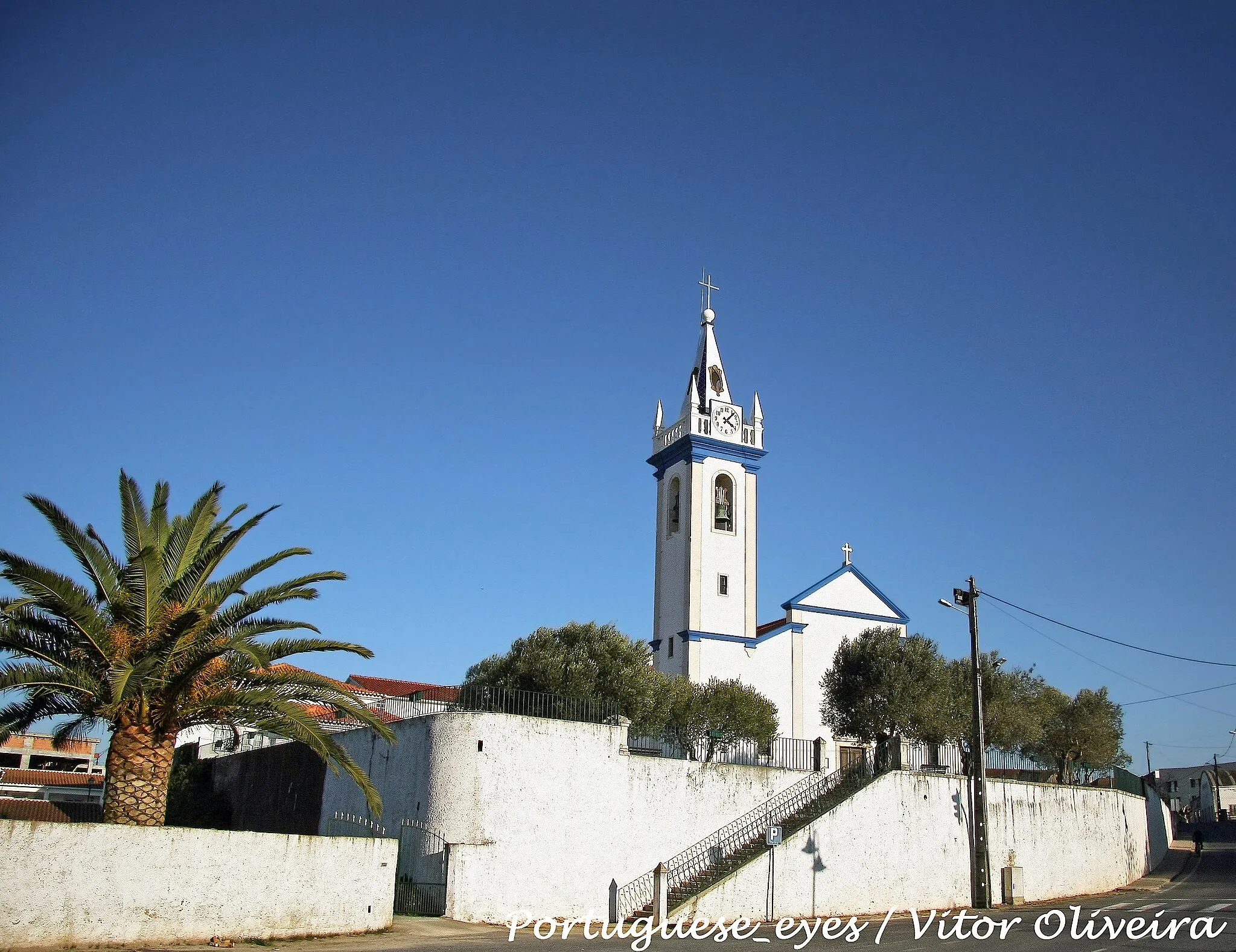 Photo showing: A Igreja Paroquial de Nossa Senhora da Luz, situa-se na entrada Oeste de A dos Cunhados e tem a fachada frontal voltada para o Parque Verde desta vila.
A actual igreja é, um testemunho precioso de uma longa história, documentando o percurso de toda a paróquia desde a sua criação até ao presente. Na sua estrutura, em três naves divididas por grossas colunas de mármore, encerra-se um precioso espólio, sobretudo em azulejaria, talha e imagens.
Na verdade, todo o corpo da igreja é percorrido por um silhar de azulejos policromados tipo tapete, datáveis de 1636, segundo os fragmentos da inscrição conservada na capela baptismal, colocada sob uma cruz feita com azulejos dispersos. Um pouco posterior, de 1656, são os azulejos da capela de S. Miguel, que incluem um painel com a Cruz e as figuras de Adão e Eva e, nos pilares, dois painéis de 2x3 cada, representando a Virgem com o Menino e uma Custódia.
A capela-mor, por seu lado, apresenta as paredes totalmente revestidas de azulejos historiados, datáveis de 1743, representando a Anunciação e a Adoração dos Pastores, sendo notável o emolduramento dos medalhões. O desenho das composições é um pouco duro, mas cheio de ritmo. As cenas, sobretudo a Adoração, são cenográficas e densamente preenchidas, seja pelos personagens aqui convocados, seja pela teatralidade de poses, bem patente na figura do arcanjo, seja já pelos elementos votivos de arquitectura e paisagem, bem como de carácter celestial (nuvens, anjos, raios de luz). Cartelas mais pequenas, sob os grande panos de azulejos descritos, apresentam outras cenas: a Purificação de Nossa Senhora no Templo, a Apresentação do Senhor, os Esponsais de São José e da Virgem e a Fuga para o Egipto. Também a sacristia apresenta um alizar em azulejos, do século XVIII, de tipo vulgar.
Regista-se também uma significativa produção de talha, sobretudo nos retábulos dos altares laterais e da capela-mor, datáveis do século XVIII, muito provavelmente resultantes da campanha de obras concluída em 1743. De notar igualmente a existência de algumas pinturas, nomeadamente a composição representando S. Miguel e as Almas, colocada na capela desta invocação (sécs. XVII-XVIII), de bastante interesse, apesar de denotar algumas repinturas.
Ao nível da estatuária, salientam-se, pela sua antiguidade, as imagens da Senhora da Luz, esculpida em pedra, datável de finais do século XVI e com decoração policromada dos séculos XVII ou XVIII, e a imagem de Santo Antão, também em pedra e policromada, quinhentista. As restantes imagens (à excepção da de S. José, Nossa Senhora de Fátima, Santo António e Sagrado Coração de Jesus), são datáveis do séc. XVIII, todas em madeira policromada e distribuídas entre a capela-mor (S. Francisco de Assis, S. Marcos) e os altares laterais (Nossa Senhora da Conceição, Menino Jesus, S. Sebastião), algumas recentemente repintadas.
Dignos de registo são também o púlpito seiscentista, assente em mísula de mármore rosa com grade de balaústres finos de madeira, as pias da água benta, da mesma época, de mármore rosa em forma de concha e a pia baptismal, em pedra lisa, provavelmente a da fundação da paróquia, nos finais do séc. XVI. Na capela-mor, relembre-se a pedra tumular assinalando a sepultura de Diogo de Teive, actualmente coberta com uma carpete de modo a proteger a respectiva inscrição.
Completam o recheio artístico da paroquial alfaias diversas, desde paramentos (com alguns têxteis seiscentistas, mas sobretudo dos sécs. XVIII e XIX) a ourivesaria, passando, entre outros, por diversos livros antigos, como o Gradual impresso em Veneza, em 1651, talvez adquirido na sequência das campanhas de embelezamento da igreja em meados da centúria.
Texto: Lucinda Rosa Pereira da Silva - João Luís Inglês Fontes praiadesantacruz.com/stcruz/religiao/igrejansraluz.htm

See where this picture was taken. [?]