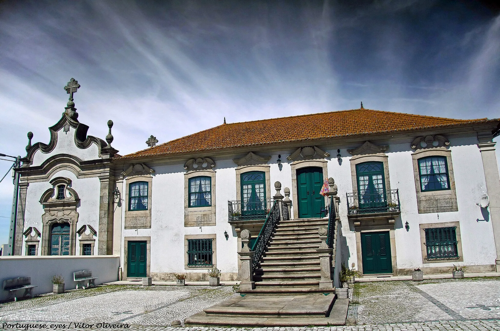 Photo showing: A freguesia de Santa Marinha de Avanca é, essencialmente, uma zona agrícola, de exploração de gado, de produção de leite (ainda que também industrial), cuja vocação lhe advém das muitas propriedades rurais que, durante séculos, a caracterizaram. Muito embora estas quintas se encontrem, hoje, bastante fragmentadas, ainda é possível perceber a sua importância e extensão, através das casas senhoriais que subsistiram (GONÇALVES, 1981). Na realidade, a presença da nobreza em Avanca remonta ao século XVI, com a fundação da Casa do Mato, mas o grande surto construtivo aconteceu nos séculos XVII e, principalmente, no século XVIII, época em que se pensa ter sido edificada a Casa do Outeiro. 
A também denominada Casa de São José do Outeiro de Paredes era sede do morgadio instituído por D. Maria Caetana de Rezende, casada com Manuel Lourenço de Sá Pereira Melo Valente, ligados por parentesco à já referida Casa do Mato (CARDOSO, 1961, p. 94). A posse da Quinta manteve-se sempre no seio desta família, até ser vendida e fragmentada, na década de 30 do século XX. O imóvel e a capela foram então adquiridos por António da Silva Valente, da Murtosa (CARDOSO, 1961, p. 95). 
Entre o conjunto de habitações senhoriais de Avanca, a casa do Outeiro é a que mais se destaca, pela imponência da sua fachada cenográfica, com ampla escadaria de acesso ao andar nobre. A frontaria é rasgada por uma série de vãos simétricos, que alternam diferentes tipologias de moldura, ao nível do segundo piso. A escadaria apresenta um amplo patamar, ladeado por pilaretes, conferindo ao conjunto algum dinamismo que, conjugado com o ritmo dos vãos, converge para o centro, evidenciando a entrada principal. 
Por sua vez, a capela encontra-se ligeiramente mais avançada em relação à fachada da Casa, mas a sua planimetria inscreve-se na mesma "tendência para o desenvolvimento num único plano" (AZEVEDO, 1988, p. 70) já observada na habitação. De reduzidas dimensões, é dominada pela composição central do portal, cujo remate, em frontão semi circular interrompido, se projecta ao nível da cimalha, que desenha o mesmo movimento. A fachada termina em frontão contracurvado, muito recortado, com fogaréus no eixo dos cunhais e cruz central. No interior, destaca-se o retábulo de talha dourada e policromada, de meados do século XVIII, que denota já o gosto rococó. Encontra-se ainda neste espaço uma campa sem identificação, com um brasão eclesiástico dos Valentes, Oliveiras e dos Resendes (GONÇALVES, 1981). 
O acesso ao conjunto habitacional é feito através de um amplo portal, com gradaria a separar o espaço privado do público, cujo desenho permite a existência de uma pequena área, fronteira à capela, quase exclusiva.

Rosário Carvalho www.patrimoniocultural.pt/pt/patrimonio/patrimonio-imovel...