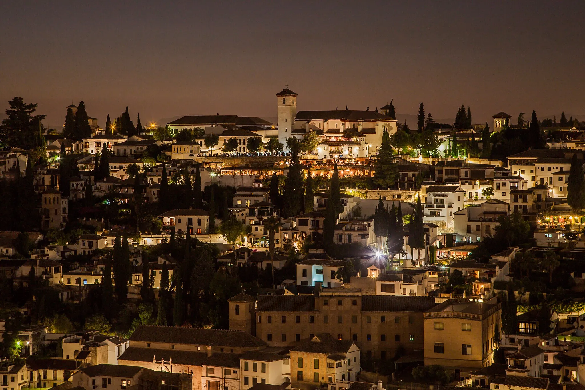 Photo showing: L'Alhambra (en arabe : الْحَمْرَاء, Al-Ḥamrā' , « la rouge », en raison de la couleur que prennent les murailles au coucher du soleil) de Grenade est un ensemble palatial, et un des monuments majeurs de l'architecture islamique et l'acropole médiévale la plus majestueuse du monde méditerranéen.
C'est avec la Grande mosquée de Cordoue le plus prestigieux témoin de la présence musulmane en Espagne du VIIIe au XVe siècle. Leurs caractères sont d'ailleurs opposés : à la sobriété grandiose du monument religieux représentatif de la première architecture islamique, s'oppose l'exubérance de la dernière manière hispano-mauresque : celle-ci s'exprime en effet dans les palais des derniers souverains nasrides, alors en pleine décadence, et qui disparaîtront bientôt lors des derniers assauts de la Reconquista.
C'est un ensemble fortifié situé sur la colline de la Sabika, qui domine la plaine et la ville de Grenade, et qui fait face au quartier populaire et pittoresque de l'Albaicin. On y aperçoit au loin les sommets enneigés de la Sierra Nevada. Parmi ces bâtiments se trouvent notamment le palais mauresque qui fait la gloire de l'Alhambra ainsi que le palais renaissant de Charles Quint et une église édifiée à la place d'une mosquée.
Patio de los Arrayanes
Le nom vient de l'arabe الْقَلْعَةُ ٱلْحَمْرَاءُ, al-Qalʻat al-Ḥamrāʼ, c'est-à-dire « le château rouge ». Il peut s'agir d'une référence à Mohammed ben Nazar, dit الأحمر, Al-'Ahmar (« le rouge », à cause de sa barbe rousse), le fondateur de la dynastie des Nasrides, qui entra à Grenade en 1238 et fonda le site, mais le toponyme semble plus ancien. Son fils Mohammed II le fortifia. Le style nasride atteint son apogée au XIVe siècle sous les rois Youssouf Ier et Mohammed V al-Ghanî, qui font édifier les parties les plus prestigieuses entre 1333 et 1354. Chaque souverain reprenait le palais de son prédécesseur et en édifiait de nouvelles parties, le modifiant à sa guise : on parle donc de palais Nasrides, au pluriel, pour cet ensemble.
Alors que presque partout dans le monde musulman les palais anciens ont disparu ou ne sont plus que des ruines, l’Alhambra possède encore deux groupes de palais du XIVe siècle. Les demeures bâties par les premiers souverains de la dynastie ont disparu et, au XVe siècle, les rois de Grenade n’ont pas eu les ressources nécessaires pour remplacer les palais subsistant aujourd’hui.
Après le règne des Nasrides de l'ombre, malgré le désir des Rois Catholiques d'effacer les traces de l'Islam des territoires entièrement reconquis par les chrétiens après la chute de Grenade en 1492, le palais mauresque était tellement superbe qu'il fut épargné et servit de résidence royale lorsque la cour passait à Grenade. Les souverains y proclament le Décret de l'Alhambra.
L'ensemble tomba ensuite en désuétude, ne faisant l'objet de restaurations qu'à l'occasion de séjours royaux.
L'Alhambra étant dès lors un des grands événements historiques, les pillards y firent leur apparition, ce que décrit Washington Irving dans ses contes.
Une action d'éclat sauva l'Alcazaba de la destruction pendant la guerre civile espagnole.
Les jardins sont à présent entretenus grâce au Patronato de La Alhambra, qui gère l'ensemble du monument et permet la visite à 7000 personnes par jour.
La gazelle est le symbole de l'Alhambra, elle est assimilable à un emblème héraldique depuis l'exploitation touristique du site. Cette image correspond à la version stylisée d'un vase décoratif retrouvée parmi les objets de l'Alhambra ; l'original se trouve dans le musée du palais de Charles Quint.
Le plan d'ensemble des Palais nasrides s'organise en de multiples salles rectangulaires, partiellement intégrées à l'enceinte, reliées entre elles par des vestibules. L'ensemble est disposé autour des deux joyaux de l'Alhambra de Grenade :
La Cour des Myrtes (Patio de los Arrayanes), au pied de la Tour de Comares et du Salon des Ambassadeurs.
La Cour des Lions (Patio de los Leones).
On peut autant parler d'un décor que d'une architecture. L'extérieur est très sobre, la décoration intérieure est foisonnante, à base des trois composantes des arts d'Islam: la calligraphie, la décoration florale stylisée, les arabesques et motifs géométriques.
La Cour des Lions est une cour rectangulaire de 35 m de long sur 20 m de large, entourée d'une galerie couverte délimitée par 124 colonnes de marbre blanc avec arcs outrepassés et riche ornementation de filigrane. Un pavillon fait saillie dans la cour à chaque extrémité, couvert d'un toit pyramidal et avec une décoration en continuité de celle de la galerie couverte. Le sol a un pavement coloré et les murs portent jusqu'à la hauteur de 1,5 m une décoration d'azulejos bleus et jaunes. Les colonnes supportant les toits et la couverture de la galerie sont espacées de manière irrégulière. Elles sont ornées de motifs végétaux variés. Au-dessus de chaque arche et au-dessus de chaque colonne sont délimités des panneaux rectangulaires décorés d'arabesques et de filigranes sculptés. Au centre de la cour se trouve la Fontaine des Lions, bassin d'albâtre supporté par douze statues de lions en marbre blanc assez grossièrement figurés mais symbolisant la puissance et le courage. Cette fontaine, d'origine judaïque, faisait partie du palais de Naghrela.
C'était le palais d'été des princes Nasrides. Ils venaient s'y rafraîchir dans les ombrages, près des bassins d'eau. Le Généralife est situé hors des murs d'enceinte, sur l'autre versant du plateau principal. Ce palais est très connu en Espagne et plusieurs poèmes ont été écrits sur ce sujet. Le nom est un dérivé de l'arabe « Jannat al-Arif  » signifiant « paradis » ou « jardins de l'architecte ».
L'abondance de l'eau dominée par les sommets enneigés de la Sierra Nevada fut pour tous ces princes issus du désert une véritable révélation. L'Alhambra et les jardins du Généralife sont les symboles les plus forts de cette domestication de l'eau qui rafraîchissait chaque cour et jardin.
The Alhambra (Arabic: الحمراء, Al-Hamra, "Red" because of the color are the walls at sunset) of Granada is a palatial complex, and one of the major monuments of Islamic architecture and the most majestic of the medieval Mediterranean world acropolis.
With the Great Mosque of Cordoba's most prestigious indicator of the Muslim presence in Spain from the eighth to the fifteenth century. Their characters are also opposites: the grand simplicity of religious monument representative of early Islamic architecture, opposes the exuberance of the last Moorish manner: it finds expression in the palaces of past rulers Nazari, then decadent, and will soon disappear in the last onslaught of the Reconquista.
It is a walled complex located on the hill of Sabika, overlooking the plain and the city of Granada, which faces the popular and picturesque area of ​​the Albaicin. One can see the distant snow-capped peaks of the Sierra Nevada. These buildings are located including the Moorish palace which is the glory of the Alhambra and the Renaissance palace of Charles V and a church built at the site of a mosque.
Patio de los Arrayanes
The name comes from the Arabic القلعة ٱلحمراء, al-Qal al-Ḥamrā', that is to say the "red castle". This may be a reference to Mohammed ben Nazar said الأحمر Al-'Ahmar ("red" because of his red beard), the founder of the Nasrid dynasty, which came to Granada in 1238 and founded the site, but the place name seems older. His son Mohammed II strengthened him. The Nasrid style reached its peak in the fourteenth century under kings Yusuf I and Mohammed V al-Ghani, who build the most prestigious parts between 1333 and 1354, each sovereign regained the palace of his predecessor and was building new parts, the modifying at will: therefore we speak of Nasrid palaces, plural, for this set.
While most parts of the Muslim world the ancient palaces have disappeared or are in ruins, yet the Alhambra palace has two groups of the fourteenth century. The houses built by the former rulers of the dynasty and disappeared in the fifteenth century, the kings of Granada did not have the resources to replace the remaining palace today.
After the reign of Nasrid shade, despite the desire of the Catholic Monarchs to erase the traces of Islam fully reconquered by the Christians after the fall of Granada in 1492 territories, the Moorish palace was so great that he was saved and used as a royal residence when the court went to Granada. The sovereigns will proclaim the decree of the Alhambra.
All fell into disuse, not subject to restorations that during royal visits.
The Alhambra is therefore one of the great historical events, looters made ​​their appearance, that Washington Irving described in his stories.
A brilliant action saved the Alcazaba from destruction during the Spanish Civil War.
The gardens are now maintained by the Patronato de la Alhambra, which manages the entire monument and gives access to 7,000 people per day.
The gazelle is the symbol of the Alhambra, it is similar to a heraldic emblem for tourist exploitation of the site. This image is a stylized decorative vase found among the objects of the Alhambra release; the original is in the museum of the palace of Charles V.
The overall layout of the Nasrid Palace is organized into multiple rectangular rooms, partially built into the enclosure, connected by hallways. The assembly is placed around the two jewels of the Alhambra in Granada
The Court of the Myrtles (Patio de los Arrayanes) at the foot of the Tower of Comares and the Salon des Ambassadeurs.
The Court of the Lions (Patio de los Leones).
We can talk as much of a decoration than an architecture. The exterior is very simple, the interior is abundant, containing the three components of Islamic art: calligraphy, stylized floral decoration, arabesques and geometric patterns.
The Court of the Lions is a rectangular court 35 m long and 20 m wide, surrounded by a covered gallery bounded by 124 white marble columns with horseshoe arches and rich ornamentation of filigree. A pavilion projects into the court at each end, covered with a pyramidal roof and decorated in line with that of the covered gallery. The floor has a colorful pavement and the walls are up to 1.5 m high decoration of blue and yellow tiles. The columns supporting the roof and cover the gallery are irregularly spaced. They are decorated with plant motifs varied. Above each arch and above each column are defined rectangular panels decorated with arabesques and carved filigree. In the center of the courtyard is the Fountain of Lions, alabaster basin supported by twelve statues of lions in white marble but rather crudely depicted symbolizing the power and courage. This fountain of Jewish descent, was in the palace of Naghrela.
It was the summer palace of the Nasrid princes. They came there to cool off in the shade, near pools of water. The Generalife is located outside the city walls, on the other side of the main board. This palace is famous in Spain and many poems have been written on this subject. The name is derived from the Arabic "Jannat al-Arif," meaning "heaven" or "garden of the architect."
The abundance of water dominated by the snowy peaks of the Sierra Nevada was for all these princes from the desert a revelation. The Alhambra and Generalife gardens are the strongest of the domestication of water that refreshed every yard and garden symbols.
قصر الحمراء (عربي: الحمراء، الحمرا، "الحمراء" بسبب لون هي الجدران عند غروب الشمس) غرناطة ومجمع فخم، واحدة من المعالم الرئيسية للعمارة الإسلامية والأكثر مهيب من القرون الوسطى الاكروبول العالم البحر الأبيض المتوسط.
مع الجامع الكبير في قرطبة مؤشر أرقى من الوجود الإسلامي في إسبانيا من الثامن إلى القرن الخامس عشر. شخصياتهم هي أيضا الأضداد: البساطة الكبرى من ممثل النصب الديني العمارة الإسلامية المبكرة، تعارض الوفرة من الطريقة مغاربي مشاركة: يجدها التعبير في قصور الحكام الماضي نظري، ثم منحط، وسوف تختفي قريبا في الهجوم الأخير من الاسترداد.
وهو مجمع مسور تقع على تلة سبيكة، ويطل على سهل ومدينة غرناطة، التي تواجه منطقة شعبية والخلابة من Albaicin. يمكن للمرء أن يرى القمم المغطاة بالثلوج بعيدة من سييرا نيفادا. وتقع هذه المباني بما في ذلك القصر المغربي الذي هو مجد قصر الحمراء وقصر عصر النهضة شارل الخامس وكنيسة بنيت في موقع مسجد.
الباحة دي لوس Arrayanes
اسم يأتي من القلعة العربية ٱلحمراء، AL-Qal الحمرا "، وهذا يعني أن" القلعة الحمراء ". قد يكون هذا وقالت في اشارة الى محمد بن نزار آل الأحمر'Ahmar ("الحمراء" بسبب لحيته الحمراء)، مؤسس سلالة بني نصر، إلى غرناطة الذي جاء في 1238 و أنشأ الموقع، ولكن اسم المكان يبدو أكبر سنا. ابنه محمد الثاني يقويه. بلغ غرار النصرية ذروته في القرن الرابع عشر تحت ملوك يوسف الأول ومحمد الخامس آل الغني، الذين يبنون الأجزاء المرموقة بين 1333 و 1354، استعادت كل سيادية القصر سلفه وكانت تبني أجزاء جديدة، و تعديل في الإرادة: وبالتالي نحن نتكلم من النصرية القصور والجمع، لهذه المجموعة.
في حين أن معظم أنحاء العالم الإسلامي القصور القديمة قد اختفت أو هي في حالة خراب، إلا أن قصر الحمراء لديه مجموعتين من القرن الرابع عشر. المنازل التي بنيت من قبل الحكام السابقين من سلالة واختفى في القرن الخامس عشر، فإن ملوك غرناطة لا تملك الموارد اللازمة لاستبدال القصر المتبقية اليوم.
بعد عهد بني نصر الظل، على الرغم من رغبة الملوك الكاثوليك لمحو آثار الإسلام معاد بالكامل من قبل المسيحيين بعد سقوط غرناطة عام 1492 الأراضي، كان القصر مغاربي كبير لدرجة أنه كان حفظها واستخدامها كأداة من الإقامة الملكية عندما ذهبت المحكمة إلى غرناطة. سوف ملوك يعلن مرسوم الحمراء.
سقطت كل داخل إهمال، لا تخضع لترميم أنه خلال زيارات المالكة.
لذا قصر الحمراء هي واحدة من الأحداث التاريخية العظيمة، جعلت اللصوص مظهرها، أن واشنطن ايرفينغ وصفها في قصصه.
والعمل الرائع حفظ القصبة من الدمار خلال الحرب الأهلية الإسبانية.
يتم الاحتفاظ الحدائق الآن من قبل باتروناتو دي لا قصر الحمراء، التي تدير النصب بأكمله ويتيح الوصول إلى 7،000 شخص يوميا.
الغزال هو رمز من قصر الحمراء، وهو مشابه إلى شارة الشعار لأغراض الاستغلال السياحي للموقع. هذه الصورة هي مزهرية الزخرفية المجردة وجدت بين الكائنات الإفراج الحمراء. النص الأصلي في متحف قصر شارل الخامس
ويتم تنظيم التخطيط الشامل للقصر النصرية في غرف مستطيلة متعددة، مبنية جزئيا في الضميمة، متصلة بواسطة الممرات. يتم وضع التجمع حول اثنين من المجوهرات من قصر الحمراء في غرناطة
المحكمة للنباتات الآس (الباحة دي لوس Arrayanes) عند سفح برج Comares وصالون السفراء.
محكمة ليونز (الباحة دي لوس ليون).
يمكننا الحديث مثل كثير من الديكور من بنية. الخارج هو بسيط جدا، والداخلية وفيرة، وتحتوي على العناصر الثلاثة للفن الإسلامي: الخط العربي والزخرفة بأسلوب منمق الأزهار، الأرابيسك وأنماط هندسية.
محكمة ليونز هي محكمة مستطيلة 35 مترا و 20 مترا، وتحيط بها معرض تغطيتها تحدها 124 أعمدة من الرخام الأبيض مع الأقواس حدوة حصان والزخرفة الغنية من الصغر. وهناك مشاريع الجناح إلى المحكمة في كل نهاية، مع تغطية سقف هرمي ومزينة بما يتماشى مع أن من معرض تغطيتها. الطابق يحتوي على الرصيف الملونة والجدران ما يصل الى 1.5 متر الديكور عال من البلاط الأزرق والأصفر. متباعدة الأعمدة تدعم سقف وتغطية معرض بشكل غير منتظم. وهي مزينة بأشكال نباتية متنوعة. فوق كل قوس وفوق كل عمود يتم تحديد وحات مستطيلة مزينة الأرابيسك ومنحوتة الصغر. في وسط الفناء هو ينبوع الليونز، حوض المرمر بدعم من اثني عشر تماثيل الأسود في الرخام الأبيض يصور بفجاجة، بل ترمز إلى القوة والشجاعة. هذا نافورة من أصل يهودي، كان في قصر Naghrela.
كان القصر الصيفي للأمراء بني نصر. جاءوا إلى هناك تهدئة في الظل قرب برك من المياه. يقع العريف خارج أسوار المدينة، وعلى الجانب الآخر من اللوحة الرئيسية. هذا القصر هو الشهير في اسبانيا وتم كتابة العديد من القصائد حول هذا الموضوع. اسم مشتق من العربية "جنات العارف،" معنى "السماء" أو "حديقة المهندس المعماري".
وكانت وفرة المياه في هذه الأندلس يهيمن عليها قمم جبال سييرا نيفادا لجميع هؤلاء الأمراء من الصحراء الوحي. حدائق قصر الحمراء والعريف هي أقوى من تدجين المياه التي استرجعت كل ساحة وحديقة حرف.
La Alhambra (en árabe: الحمراء, Al-Hamra, "rojo" por el color son las paredes al atardecer) de Granada es un complejo palaciego, y uno de los principales monumentos de la arquitectura islámica y el más majestuoso de los medievales acrópolis mundo mediterráneo.
Con la Gran Mezquita de Córdoba del más prestigioso indicador de la presencia musulmana en España desde el siglo VIII al XV. Sus personajes son también opuestos: la gran simplicidad de monumento religioso representativo de la arquitectura islámica, se opone a la exuberancia de la última manera morisca: que se expresa en los palacios de los gobernantes del pasado Nazari, entonces decadente, y está próximo a desaparecer en el último ataque de la Reconquista.
Es un complejo amurallado situado en la colina de la Sabika, que domina la llanura y la ciudad de Granada, que da a la zona popular y pintoresco barrio del Albaicín. Uno puede ver los picos nevados distantes de la Sierra Nevada. Estos edificios se encuentran incluidos el palacio árabe que es la gloria de la Alhambra y el palacio renacentista de Carlos V y una iglesia construida en el sitio de una mezquita.
Patio de los Arrayanes
El nombre proviene del árabe القلعة ٱلحمراء, al-Qal al-Hamra ', es decir, el "castillo rojo". Esto puede ser una referencia a Mohammed ben Nazar dijo الأحمر Al-'Ahmar ("rojo" debido a su barba roja), el fundador de la dinastía nazarí, que llegó a Granada en 1238 y fundada el sitio, pero el nombre del lugar parece viejo. Su hijo Mohamed II lo fortaleció. El estilo nazarí alcanzó su apogeo en el siglo XIV bajo reyes Yusuf I y Mohamed V al-Ghani, que construyen las partes más prestigiosas entre 1333 y 1354, cada soberano recuperó el palacio de su predecesor y estaba construyendo nuevas piezas, los modificar a voluntad: por lo tanto, hablamos de los palacios nazaríes, en plural, para este conjunto.
Si bien la mayor parte del mundo musulmán los antiguos palacios han desaparecido o están en ruinas, sin embargo, el palacio de la Alhambra tiene dos grupos del siglo XIV. Las casas construidas por los antiguos gobernantes de la dinastía y desapareció en el siglo XV, los reyes de Granada no tenían los recursos para reemplazar el palacio que queda hoy en día.
Después del reinado nazarí de sombra, a pesar del deseo de los Reyes Católicos para borrar las huellas del Islam totalmente reconquistada por los cristianos después de la caída de Granada en 1492 territorios, el palacio árabe era tan grande que era guardado y utilizado como residencia real cuando la corte fue a Granada. Los soberanos proclamarán el decreto de la Alhambra.
Todos cayeron en desuso, que no están sujetos a las restauraciones que durante las visitas reales.
Por ello, la Alhambra es uno de los grandes acontecimientos históricos, los saqueadores hicieron su aparición, que Washington Irving describió en sus historias.
Una brillante acción salvó la Alcazaba de la destrucción durante la Guerra Civil española.
Los jardines están mantenidos por el Patronato de la Alhambra, que gestiona todo el monumento y le da acceso a 7.000 personas por día.
La gacela es el símbolo de la Alhambra, que es similar a un emblema heráldico de la explotación turística del lugar. Esta imagen es un jarrón decorativo estilizado encontrado entre los objetos de la liberación Alhambra; el original está en el museo del palacio de Carlos V.
El diseño general del palacio nazarí está organizado en varias habitaciones rectangulares, parcialmente construidas en el recinto, conectados por pasillos. El conjunto se coloca alrededor de las dos joyas de la Alhambra de Granada
El Patio de los Arrayanes (Patio de los Arrayanes) a los pies de la Torre de Comares y el Salon des Ambassadeurs.
El Patio de los Leones (Patio de los Leones).
Podemos hablar tanto de una decoración de una arquitectura. El exterior es muy simple, el interior es abundante, que contiene los tres componentes del arte islámico: la caligrafía, estilizada decoración floral, arabescos y motivos geométricos.
El Patio de los Leones es un campo rectangular de 35 m de largo y 20 m de ancho, rodeado por una galería cubierta limitada por 124 columnas de mármol blanco con arcos de herradura y rica ornamentación de filigrana. Un pavilion proyecta en la corte en cada extremo, cubierto con un techo piramidal y decoradas en consonancia con el de la galería cubierta. El piso tiene un pavimento de colores y las paredes son de hasta 1,5 m de altura decoración de azulejos azules y amarillos. Las columnas que sostienen el techo y cubrir la galería están irregularmente espaciados. Están decoradas con motivos vegetales variados. Por encima de cada arco y encima de cada columna se definen paneles rectangulares decorados con arabescos y filigranas tallada. En el centro del patio se encuentra la Fuente de los Leones, cuenca de alabastro con el apoyo de doce estatuas de leones de mármol blanco, sino más bien crudamente representado que simboliza el poder y la valentía. Esta fuente de origen judío, estaba en el palacio de Naghrela.
Era el palacio de verano de los príncipes nazaríes. Llegaron allí para refrescarse en la sombra, cerca de charcos de agua. El Generalife se encuentra fuera de las murallas de la ciudad, al otro lado de la placa principal. Este palacio es famoso en España y muchos poemas se han escrito sobre este tema. El nombre se deriva del árabe "Jannat al-Arif," que significa "cielo" o "jardín del arquitecto."
La abundancia de agua dominado por los picos nevados de la Sierra Nevada era para todos estos príncipes del desierto una revelación. Los jardines de la Alhambra y el Generalife son los más fuertes de la domesticación de agua que refresca cada patio y jardín símbolos.
阿罕布拉（阿拉伯语：“红”的颜色，因为الحمراء，哈姆拉，是在日落的墙壁）格拉纳达是富丽堂皇的复杂，伊斯兰建筑的主要古迹之一而最雄伟的中世纪地中海世界的雅典卫城。
随着大清真寺科尔多瓦的最负盛名的西班牙穆斯林存在的第八至十五世纪的指标。他们的性格也相反：早期伊斯兰建筑的宗教纪念碑代表的宏伟简洁，反对的最后一个摩尔人的方式繁荣：找到过去统治者的宫殿式纳扎里，那么颓废，并很快消失在收复失地运动的最后冲击。
这是一个复杂的围墙位于Sabika的山上，俯瞰着平原和城市格拉纳达，它面临的阿尔巴辛的流行和风景如画的地区。人们可以看到内华达远处白雪皑皑的山峰。这些建筑都位于其中包括摩尔人宫殿是阿罕布拉和查理五世的文艺复兴时期的宫殿和建在一座清真寺的网站教会的荣耀。
天井德洛斯香桃木
这个名字来源于阿拉伯语القلعةٱلحمراء，AL-QAL哈姆拉“，即”红色城堡“。这可能是一个参考穆罕默德·本·纳扎尔说الأحمر铝'Ahmar（“红”，因为他的红胡子），该Nasrid王朝的奠基人，其来到格拉纳达在1238创办该网站，但这个地方的名字似乎更老。他的儿子穆罕默德二世加强了他。该Nasrid风格达到了顶峰下的国王优素福I和穆罕默德五世人 - 加尼，谁1333和1354年之间建造的最负盛名的地方在14世纪，每个主权收复了他的前任的宫殿，并建立新的部分，修改的意愿。所以我们讲Nasrid宫殿，多元，对于这一套。
而穆斯林世界大部分地区的古代宫殿已经消失或正在废墟中，但阿罕布拉宫有两组十四世纪。由王朝的统治者前建成并消失在十五世纪的房子，格拉纳达国王没有足够的资源，今天替换剩余的宫殿。
Nasrid遮阳的统治，尽管天主教君主抹去了伊斯兰教的格拉纳达于1492年地区沦陷后的基督徒完全重新征服的痕迹的愿望后，摩尔人宫殿是如此之大，他是保存并作为皇家住所时，法院去格拉纳达。君主将宣布阿罕布拉法令。
所有落入废弃，不受修复，在国王出访。
因此，阿罕布拉是伟大的历史事件之一，抢劫他们的露面，华盛顿欧文在他的故事中描述。
辉煌的行动挽救了阿尔卡萨瓦从毁灭西班牙内战期间。
花园现在由PATRONATO德拉阿罕布拉，负责管理整个纪念碑和可访问每天7000人维持。
瞪羚是阿罕布拉的象征，它类似于一个纹章徽为网站的旅游开发。此图片是阿罕布拉释放的对象中发现了一个程式化的花瓶;原来是在查理五世宫殿博物馆
在Nasrid宫的整体布局分为多个矩形的房间，部分内置于机箱，由走廊相连。该组件被放置围绕在格拉纳达的阿罕布拉两个珠宝
在桃金娘中庭（天井德洛斯香桃木）在Comares塔和沙龙des Ambassadeurs酒店的脚下。
狮子法院（天井德洛斯利昂）。
我们可以谈论尽可能多的装饰不是建筑。它的外观很简单，内饰丰富，含有伊斯兰艺术的三个组成部分：书法，程式化的花卉装饰，花纹和几何图案。
狮子法院是一个长方形场地长35米和宽20米处，通过有盖长廊与马蹄形拱门和华而不实的丰富的纹饰124的白色大理石柱为界包围。亭子项目进入法庭的两端，覆盖着金字塔形屋顶和装饰线与覆盖的画廊。地板上有一个丰富多彩的路面和墙壁是高达1.5米，高装饰蓝色和黄色的瓷砖。支撑屋顶，覆盖画廊的列间隔不规则。他们都装饰着不同的植物图案。上述每个拱和每一列定义如上饰以花纹和雕刻镂空雕花长方形板。在院子的中心是狮子喷泉，雪花盆白色大理石支撑12尊石狮，而是粗暴地描绘象征着力量和勇气。犹太血统这个喷泉，是Naghrela的宫殿。
这是Nasrid王子的夏宫。他们来到那里，在阴凉处冷却一下，水池附近。该Generalife位于城墙外，在主板的另一侧。这座宫殿是著名的西班牙和许多诗歌都描写了这个问题。这个名字是从阿拉伯语派生出来的“Jannat AL-阿里夫”，意思是“天堂”或“建筑师的花园。”
水在这个安达卢西亚丰富的内华达山脉的雪峰支配是来自沙漠的一个启示，这些诸侯。阿罕布拉和Generalife花园是最强的水驯化的刷新每院子和花园的符号。
La Alhambra (in arabo: الحمراء, Al-Hamra, "Rosso" per via del colore sono le pareti al tramonto) di Granada è un complesso palaziale, e uno dei più importanti monumenti di architettura islamica e la più maestosa delle medievali dell'acropoli mondo mediterraneo.
Con la Grande Moschea di Cordoba del più prestigioso indicatore della presenza musulmana in Spagna, dall'ottavo al quindicesimo secolo. I loro personaggi sono anche opposti: la grande semplicità del rappresentante monumento religioso di architettura islamica, si oppone l'esuberanza dell'ultima modo moresco: essa trova espressione nei palazzi dei sovrani del passato Nazari, poi decadente, e presto scomparirà l'ultimo assalto della Reconquista.
Si tratta di un complesso recintato situato sulla collina del Sabika, domina la pianura e la città di Granada, che si affaccia sulla famosa e pittoresca zona di Albaicin. Si possono vedere le cime innevate lontane della Sierra Nevada. Questi edifici sono situati tra cui il palazzo moresco che è la gloria della Alhambra e il palazzo rinascimentale di Carlo V e di una chiesa costruita sul sito di una moschea.
Patio de los Arrayanes
Il nome deriva dall'arabo القلعة ٱلحمراء, al-Qal al-Hamra ', vale a dire il "castello rosso". Questo può essere un riferimento a Mohammed ben Nazar ha detto الأحمر Al-'Ahmar ("rosso" a causa della sua barba rossa), il fondatore della dinastia Nasridi, che è venuto a Granada nel 1238 e fondato il sito, ma il toponimo sembra più vecchio. Suo figlio Maometto II lo ha rafforzato. Lo stile Nasridi raggiunto il suo picco nel XIV secolo sotto i re Yusuf I e Mohammed V al-Ghani, che costruiscono i pezzi più prestigiosi tra il 1333 e il 1354, ogni sovrano riacquistato il palazzo del suo predecessore e stava costruendo nuove parti, i modificando a volontà: quindi parliamo di palazzi Nasridi, plurale, per questo set.
Mentre la maggior parte del mondo musulmano gli antichi palazzi sono scomparsi o sono in rovina, ma il palazzo dell'Alhambra ha due gruppi del XIV secolo. Le case costruite dagli ex governanti della dinastia e scomparsi nel XV secolo, i re di Granada non hanno le risorse per sostituire il palazzo rimanente oggi.
Dopo il regno dei Nasridi ombra, nonostante la volontà dei Re Cattolici per cancellare le tracce dell'Islam pienamente riconquistata dai cristiani dopo la caduta di Granada nel 1492 i territori, il palazzo moresco era così grande che era salvato e utilizzato come residenza reale quando la corte è andato a Granada. I sovrani si proclamare il decreto di Alhambra.
Tutti caddero in disuso, non soggetta a restauri che durante le visite reali.
L'Alhambra è quindi uno dei grandi eventi storici, i saccheggiatori hanno fatto la loro comparsa, che Washington Irving descritto nei suoi racconti.
Un'azione brillante salvato la Alcazaba dalla distruzione durante la guerra civile spagnola.
I giardini sono ora gestiti dal Patronato de la Alhambra, che gestisce l'intero monumento e dà accesso a 7000 persone al giorno.
La gazzella è il simbolo della Alhambra, è simile a un emblema araldico per la valorizzazione turistica del sito. Questa immagine è un vaso decorativo stilizzato trovato tra gli oggetti del rilascio Alhambra; l'originale è nel museo del palazzo di Carlo V.
Il layout complessivo del Palazzo Nasridi è organizzato in più stanze rettangolari, parzialmente integrato nel recinto, collegati da corridoi. L'assemblea è collocato intorno ai due gioielli della Alhambra di Granada
La Corte dei mirti (Patio de los Arrayanes), ai piedi della Torre di Comares e il Salon des Ambassadeurs.
La Corte dei Leoni (Patio de los Leones).
Possiamo parlare tanto di una decorazione di una architettura. L'esterno è molto semplice, l'interno è abbondante, contenente i tre componenti di arte islamica: calligrafia, decorazione floreale stilizzata, arabeschi e motivi geometrici.
La Corte dei Leoni è una corte rettangolare lunga 35 metri e larga 20 m, circondato da una galleria coperta delimitata da 124 colonne di marmo bianco con archi a ferro di cavallo e ricchi ornamenti di filigrana. Un padiglione progetti in tribunale ad ogni estremità, coperti con un tetto piramidale e decorate in linea con quella della galleria coperta. Il piano ha un pavimento colorato e le pareti sono fino a 1,5 m di altezza decorazione di piastrelle blu e giallo. Le colonne che sostengono il tetto e coprire la galleria sono irregolarmente distanziati. Sono decorate con motivi vegetali varia. Sopra ogni arco e sopra ogni colonna vengono definiti pannelli rettangolari decorati con arabeschi in filigrana e intagliato. Al centro del cortile è la Fontana dei Leoni, bacino alabastro sostenuto da dodici statue di leoni in marmo bianco, ma piuttosto rozzamente dipinto che simboleggia la potenza e coraggio. Questa fontana di origine ebraica, era nel palazzo di Naghrela.
Era la residenza estiva dei principi Nasridi. Sono venuti lì per rinfrescarsi all'ombra, nei pressi di pozze d'acqua. Il Generalife è situato al di fuori delle mura della città, sul lato opposto della scheda principale. Questo palazzo è famoso in Spagna e in molte poesie sono state scritte su questo argomento. Il nome deriva dall'arabo "Jannat al-Arif," che significa "cielo" o "giardino dell'architetto."
L'abbondanza di acqua dominata dalle cime innevate della Sierra Nevada è stato per tutti questi principi dal deserto una rivelazione. I giardini di Alhambra e Generalife sono i più forti della domesticazione di acqua che aggiornato ogni cortile e giardino simboli.
アルハンブラ（アラビア語：الحمراء、アル·ハムラ、なぜなら色の「赤」は日没で壁である）グラナダは宮殿複雑であり、イスラム建築の主要なモニュメントの一つ中世地中海世界のアクロポリスの中で最も壮大な。
第八からスペインのイスラム教徒の存在のコルドバで最も有名な指標の大モスクでは、15世紀に。自分のキャラクターも正反対である：初期のイスラム建築の宗教的なモニュメントを代表する壮大なシンプルさは、最後のムーア人の様子の活気に反対：それは過去の支配者の宮殿内での発現を検出したNazari、その後退廃、すぐにレコンキスタの最後の猛攻に表示されなくなります。
それは、プレーンとアルバイシンの人気があり、美しいエリアに直面しているグラナダの街を見下ろすSabikaの丘の上に壁に複雑です。一つは、シエラネバダの遠い雪に覆われたピークを見ることができます。これらの建物は、アルハンブラ宮殿とチャールズVのルネサンス宮殿やモスクの敷地に建てられた教会の栄光であるムーア人の宮殿など、配置されています。
パティオ·デ·ロス·アラジャネス
名前はアラビア語القلعةٱلحمراء、アル·アル·ハムラQAL」から来ている、それは「赤い城"と言うことです。これはモハメッドベンナザールへの参照がالأحمر言ったかもしれアル'Ahmar（なぜなら彼の赤いひげの「赤」）、1238年にグラナダに来たナスル朝王朝の創始者、およびサイトを設立したが、地名は、古いようです。彼の息子モハメッドIIは彼を強化した。ナスル朝のスタイルは、それぞれの主権は、彼の前任者の宮殿を取り戻し、新しいパーツを構築し、1333年と1354年の間で最も権威のある部品を構築王ユスフIとモハメッド·アル·気前がいい、下の14世紀にピークに達し、意のままに変更すること：それゆえ私たちは、このセットのために、複数のナスル朝宮殿、話す。
イスラム世界のほとんどの地域ながら、古代の宮殿は消えてしまったかの遺跡にあり、まだアルハンブラ宮殿は、14世紀の2つのグループを持っています。 15世紀に王朝の旧支配者によって構築され、姿を消した家は、グラナダの王たちは、今日、残りの宮殿を交換するためのリソースを持っていませんでした。
ナスル朝シェードの治世の後、完全に1492地域でグラナダ陥落後にキリスト教徒reconqueredイスラムの痕跡を消去するカトリック両王の願いにもかかわらず、ムーア人の宮殿は、彼があったことをとても素晴らしかった保存され、裁判所はグラナダに行ったときの王宮として使用。ソブリンはアルハンブラ令を宣言します。
すべては、その王室の訪問の間、修復の対象ではない、使われなくなりました。
アルハンブラ、したがって偉大な歴史的なイベントの一つであり、略奪者は、ワシントンアーヴィングが彼の話で説明していることを、彼らの顔を見せた。
華麗なアクションがスペイン内戦中に破壊からアルカサバを救った。
庭園は現在、全体碑を管理し、一日あたり7,000人へのアクセスを提供しますPatronatoデラアルハンブラ、によって維持されている。
ガゼルはアルハンブラの象徴であり、それは、サイトの観光開発のための紋章のエンブレムに似ています。この画像はアルハンブラのリリースのオブジェクトの間で発見定型化された装飾的な花瓶です。オリジナルのチャールズVの宮殿の博物館である
ナスル朝宮殿の全体的なレイアウトは廊下で接続され、部分的に囲いに組み込まれた複数の長方形の部屋、、で構成されています。アセンブリは、グラナダのアルハンブラ宮殿の2宝石の周りに配置されている
コマレスの塔、サロンデアンバサダーの麓Myrtles裁判所（パティオ·デ·ロス·アラジャネス）。
ライオンズの裁判所（パティオ·デ·ロス·ライオン）。
私たちは、アーキテクチャよりも装飾をできるだけ多く話すことができます。書道、様式化された花の装飾、アラベスクや幾何学模様：エクステリア、インテリアは、イスラム美術の3つの成分を含む、豊富な、非常に簡単です。
ライオンズの裁判所は、馬蹄アーチとフィリグリーの豊かな装飾を持つ124白い大理石の柱で囲まれた覆われ、ギャラリーに囲まれた、長方形の35メートルの長いコート、20メートル幅である。各端に裁判所へのパビリオンプロジェクト、ピラミッド型の屋根で覆われ、覆われたギャラリーのそれとラインで装飾。床はカラフルな舗装があり、壁は青と黄色のタイルの1.5メートルの高デコレーション次第である。ギャラリーを屋根を支え、カバーの列が不規則に配置されている。これらは変動し、植物のモチーフが施されています。各アーチの上に、各列の上にアラベスクと刻まれた細工で飾られた長方形のパネルを定義されています。中庭の中央には、ライオンズの噴水、アラバスター流域白大理石でライオンの彫像12でサポートされているのではなく粗雑力と勇気を象徴する描写がある。ユダヤ系のこの噴水は、Naghrelaの宮殿にいた。
それは、ナスル朝の王子の夏の宮殿であった​​。彼らは水のプールの近くで、日陰で涼んあるようになった。ヘネラリフェは、メインボードの反対側に、城壁の外に位置しています。この宮殿はスペインで有名で、多くの詩はこの主題について書かれている。名前は「天」または意味する「Jannatアル·アリフ"アラビア語から派生した「建築家の庭に。"
シエラネバダの雪のピークによって支配このアンダルシアの水の存在量は、砂漠の啓示から、これらすべての諸侯のためだった。アルハンブラ宮殿とヘネラリフェ庭園は、すべての庭と庭園のシンボルをリフレッシュさの水の家畜化の最強です。
A Alhambra (em árabe: الحمراء, Al-Hamra, "Red" por causa da cor são as paredes, pôr do sol) de Granada é um complexo palaciano, e um dos principais monumentos da arquitetura islâmica eo mais majestoso dos medievais acrópole mundo mediterrâneo.
Com a Grande Mesquita de Córdoba de maior prestígio indicador da presença muçulmana na Espanha a partir do oitavo ao século XV. Seus personagens também são opostos: o grande simplicidade de monumento religioso representante da arquitetura islâmica cedo, opõe-se à exuberância da última forma moura: ela encontra expressão nos palácios dos governantes passados Nazari, então decadente, e irá desaparecer em breve no último ataque da Reconquista.
É um complexo murado localizado na colina de Sabika, com vista para a planície e da cidade de Granada, que enfrenta a área popular e pitoresca do Albaicín. Pode-se ver os picos nevados distantes da Sierra Nevada. Estes prédios estão localizados, incluindo o palácio mouro que é a glória da Alhambra e do palácio renascentista de Carlos V e uma igreja construída no local de uma mesquita.
Patio de los Arrayanes
O nome vem do árabe القلعة ٱلحمراء, al-Qal al-Hamra ', isto é, o "castelo vermelho". Isto pode ser uma referência a Mohammed ben Nazar disse الأحمر Al-'Ahmar ("vermelho" por causa de sua barba vermelha), o fundador da dinastia Nasrid, que chegou a Granada em 1238 e fundou o site, mas o nome da cidade parece mais velho. Seu filho Mohammed II reforçou ele. O estilo Nasrida atingiu o seu auge no século XIV, sob os reis Yusuf I e Mohammed V al-Ghani, que constroem as peças de maior prestígio entre 1333 e 1354, cada soberano recuperou o palácio de seu antecessor e foi a construção de novas peças, os modificar à vontade: por isso falamos de Nasrid palácios, plural, para este conjunto.
Enquanto a maioria das partes do mundo muçulmano os palácios antigos desapareceram ou estão em ruínas, mas o palácio Alhambra tem dois grupos de do século XIV. As casas construídas pelos antigos governantes da dinastia e desapareceram no século XV, os reis de Granada não têm os recursos para substituir o palácio que permanecem hoje.
Após o reinado de Nasrid sombra, apesar do desejo dos Reis Católicos para apagar os traços do Islã totalmente reconquistada pelos cristãos após a queda de Granada em 1492 territórios, o palácio mouro era tão grande que ele foi guardados e utilizados como residência real, quando o tribunal foi a Granada. Os soberanos irá proclamar o decreto do Alhambra.
Tudo caiu em desuso, não sujeito a restaurações que durante as visitas reais.
A Alhambra é, portanto, um dos grandes acontecimentos históricos, saqueadores fez sua aparição, que Washington Irving descreveu em suas histórias.
A ação brilhante salvou a Alcazaba de destruição durante a Guerra Civil Espanhola.
Os jardins são agora mantida pelo Patronato de la Alhambra, que gerencia todo o monumento e dá acesso a 7.000 pessoas por dia.
A gazela é o símbolo da Alhambra, é semelhante a um emblema heráldico para a exploração turística do local. Esta imagem é um vaso decorativo estilizado encontrado entre os objetos da liberação Alhambra; o original está no museu do palácio de Carlos V.
O layout geral do Palácio Nasrid está organizado em várias salas retangulares, construído parcialmente dentro do gabinete, ligadas por corredores. O conjunto é colocado em torno das duas jóias da Alhambra, em Granada
O Tribunal de Murta (Patio de los Arrayanes) ao pé da Torre de Comares eo Salon des Ambassadeurs.
O corte dos leões (Patio de los Leones).
Podemos falar tanto de uma decoração de uma arquitetura. O exterior é muito simples, o interior é abundante, contendo os três componentes da arte islâmica: caligrafia, decoração floral estilizado, arabescos e formas geométricas.
O corte dos leões é uma quadra retangular de 35 m de comprimento e 20 m de largura, rodeado por uma galeria coberta delimitada por 124 colunas de mármore branco com arcos de ferradura e rica ornamentação de filigrana. Um pavilhão projeta-se o corte em cada extremidade, cobertos com um telhado piramidal e decorados de acordo com a da galeria coberta. O chão tem um pavimento colorido e as paredes são de até 1,5 m de altura decoração de azulejos azuis e amarelos. As colunas que sustentam o telhado e cobrem a galeria são espaçados irregularmente. Eles são decorados com motivos vegetais variados. Acima de cada arco e acima de cada coluna são definidas painéis retangulares decoradas com arabescos e filigranas esculpidas. No centro do pátio é a Fonte dos Leões, a bacia de alabastro suportado por doze estátuas de leões em mármore branco, mas sim cruamente retratado simbolizando o poder e coragem. Esta fonte de ascendência judaica, estava no palácio de Naghrela.
Era o palácio de verão dos príncipes Nazaris. Eles vieram lá para se refrescar na sombra, perto de poças de água. O Generalife está localizado fora das muralhas da cidade, do outro lado da placa principal. Este palácio é famoso na Espanha e em muitos poemas foram escritos sobre este assunto. O nome é derivado do árabe "al-Arif Jannat", que significa "céu" ou "jardim do arquiteto".
A abundância de água dominada pelos picos nevados da Sierra Nevada foi para todos estes príncipes do deserto uma revelação. Os jardins de Alhambra e Generalife são os mais fortes da domesticação da água que atualizada a cada quintal e jardim símbolos.
Альгамбра (Арабский: الحمراء, Аль-Хамра, "Красный" из-за цвета являются стены на закате) Гранада роскошный комплекс, и одним из главных памятников исламской архитектуры и самый величественный из средневековых средиземноморских мировых акрополя.
С Великая мечеть Кордовы самых престижных показателя присутствия мусульман в Испании от восьмого до пятнадцатого века. Их персонажи также противоположности: великий простота религиозный памятник представителя раннего исламского зодчества, выступает против изобилие последнего мавританского образом: она находит свое выражение во дворцах последние правителей Назари, то декадентской, и скоро исчезнет в прошлом натиском Реконкисты.
Это стеной комплекс, расположенный на холме Sabika, с видом на равнину и город Гранада, которая стоит перед популярный и живописном районе Albaicin. Видно далекие заснеженные пики Сьерра-Невада. Эти здания расположены в том числе дворца мавританского который слава Альгамбры и ренессансного дворца Карла V и церкви, построенной на месте мечети.
Патио-де-лос-Arrayanes
Название происходит от арабского القلعة ٱلحمراء, аль-Qal аль-Хамра ", то есть" красный замок ". Это может быть ссылка на Мухаммеда бен Назар сказал الأحمر Аль-'Ahmar ("красный", потому что его рыжей бородой), основатель династии Насридов, который приехал в Гранаду в 1238 и основал сайт, но название кажется старше. Его сын Мохаммед II укрепил его. Стиль Nasrid достигла своего пика в четырнадцатом веке под королей Юсуф I и Мухаммед V аль-Гани, которые строят самые престижные части между 1333 и 1354, каждое суверенное восстановил дворец своего предшественника и строил новые детали, в изменения по желанию: поэтому мы говорим о Nasrid дворцов, множества, для этого набора.
В то время как большинство частях мусульманского мира древние дворцы исчезли или находятся в руинах, пока дворец Альгамбра имеет две группы в четырнадцатом веке. Дома, построенные по бывших правителей династии и исчезли в пятнадцатом веке, цари Гранада не имеют ресурсов, чтобы заменить оставшиеся дворец сегодня.
После правления Насридов тени, несмотря на желание католических монархов, чтобы стереть следы ислама полностью отвоевали христианами после падения Гранады в 1492 территорий, дворец в мавританском был настолько велик, что он был сохранить и использовать в качестве королевской резиденции, когда суд пошел в Гранаду. Государи возвещу Альгамбра.
Все вышел из употребления, не подлежат реставрации, что во время королевских визитов.
Поэтому Альгамбра является одним из великих исторических событий, мародеры появились, что Вашингтон Ирвинг описал в своих рассказах.
Блестящий действия спас Алькасаба от разрушения во время гражданской войны в Испании.
Сады в настоящее время поддерживается Patronato де-ла-Альгамбра, которая управляет всей памятник и дает доступ к 7000 человек в день.
Газель является символом Альгамбра, он похож на геральдические эмблемы для туристической эксплуатации сайта. Это изображение является стилизованный декоративные вазы найдены среди объектов выпуском Альгамбра;Оригинал находится в музее дворца Карла V.
Общая компоновка Насридов дворца организована в нескольких прямоугольных комнат, частично встроенные в корпуса, соединенных коридорами. Сборка помещается около двух жемчужин Альгамбра в Гранаде
Суд из Myrtles (Патио-де-лос-Arrayanes) у подножия башни Комарес и Салоне Ambassadeurs.
Суд о Львове (Патио-де-лос-Леонес).
Мы можем говорить, как много украшения, чем архитектуры. Внешность очень простой, интерьер в изобилии, содержащий три компонента исламского искусства: каллиграфия, стилизованные цветочные украшения, арабесками и геометрическими узорами.
Суд львов представляет собой прямоугольное суд длиной 35 м и шириной 20 м, окруженный крытой галереей, ограниченной 124 белых мраморных колонн с подковы арок и богатым орнаментом из филиграни. Павильон выступающий в суде на каждом конце, покрытые пирамидальной крышей и оформленные в соответствии с, что из крытой галереи. Пол имеет красочный тротуар и стены до 1,5 м в высоту украшения синие и желтые плитки. Колонны, поддерживающие крышу и покрыть галерею неравномерно расположены. Они украшены растительными мотивами варьировались. Над каждой аркой и выше каждого столбца определены прямоугольные панели, украшенные арабесками и резной филигранью. В центре двора находится фонтан Львов, алебастр бассейна поддерживается двенадцати статуй львов из белого мрамора, но довольно грубо изображены символизирующий силу и мужество. Этот фонтан еврейского происхождения, был во дворце Naghrela.
Это был летний дворец князей Nasrid. Они пришли туда, чтобы остыть в тени, рядом лужам.Хенералифе находится за пределами городских стен, на другой стороне основной платы. Этот дворец известен в Испании и многие стихи были написаны на эту тему. Название происходит от арабского "​​Жаннат аль-Ариф", что означает "небо" или "сад архитектора».

Обилие воды в этом Андалусии преобладают заснеженных вершин Сьерра-Невада был для всех этих князей из пустыни откровение. Альгамбра и сады Хенералифе являются самыми сильными из одомашнивания воды, что обновление каждые двор и сад символов.