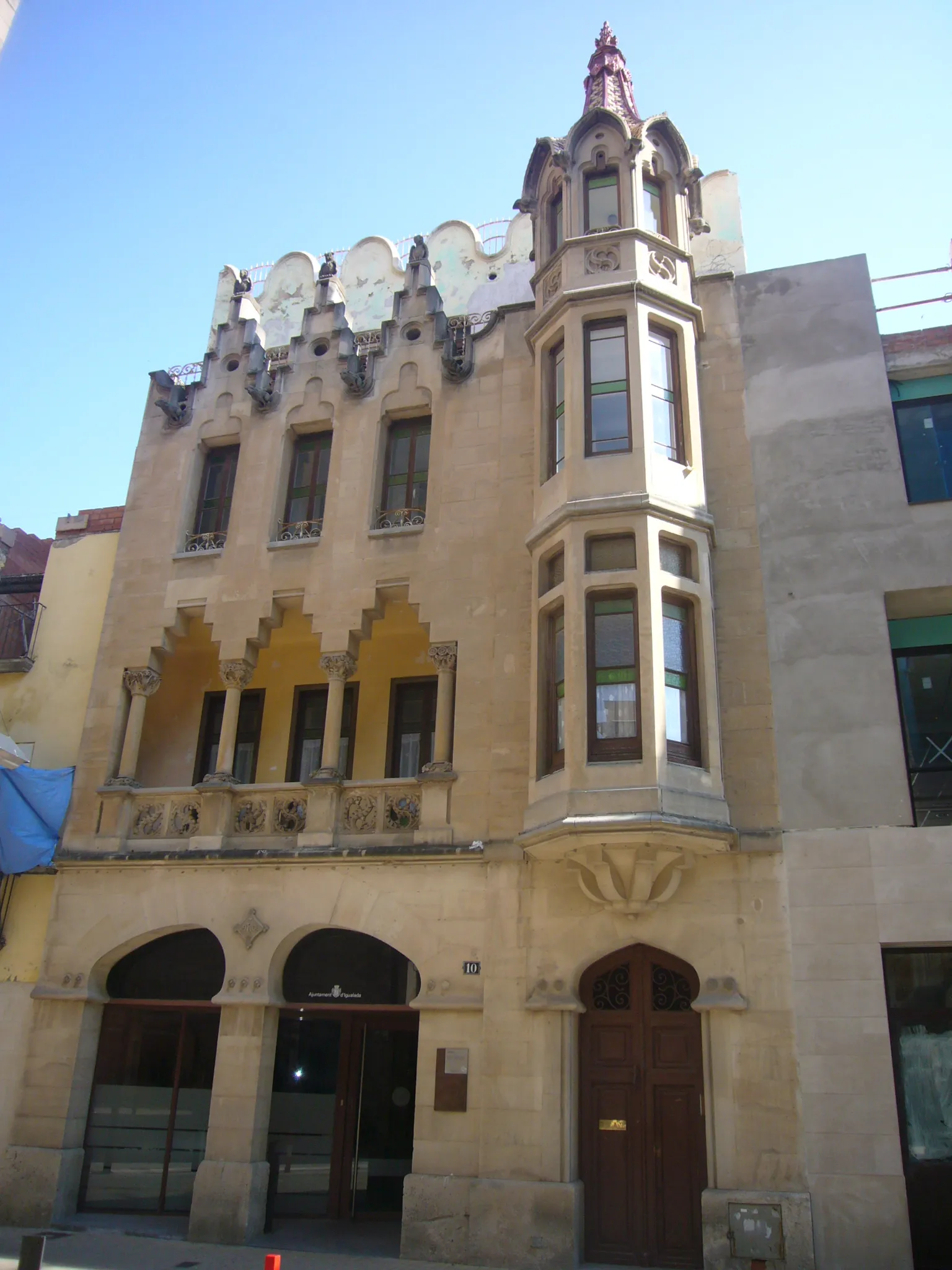 Photo showing: CAL RATÉS: Edifici d’estatges i planta comercial al número 8-10 del carrer de santa Maria (Igualada), construït l'any 1908 pels arquitectes Isidre Gili Moncunill i Pau Salvat Espasa. La façana presenta una tribuna coronada per un pinacle de ceràmica vidriada de tons vermellosos i grocs. La part comercial de la planta baixa –avui ocupada per oficines municipals- presenta una porta geminada d’arcs rampants. La porta d’accés als habitatges presenta una adaptació modernista de l’arc de ferradura. Al primer pis hi ha una galeria d’arcs esglaonats. Rematen el tercer pis unes falses gàrgoles entre merlets, coronats per figures de mussols. Si a la façana principal l’element constructiu és la pedra, a la part posterior, que dóna a la plaça sant Miquel, l’element emprat és el maó vist, en combinació amb la ceràmica vidriada de color verd, utilitzada en els múltiples pinacles que coronen l’edifici. A l’interior dels estatges també hi trobem elements típicament modernistes, com son les llars de foc, bastides amb marbres. Data del projecte: Octubre 1908.Propietari promotor de l'obra: Marià Rates. Ofici del propietari promotor: Comerç. Plànols: Lligall 1908 - Lletra C.