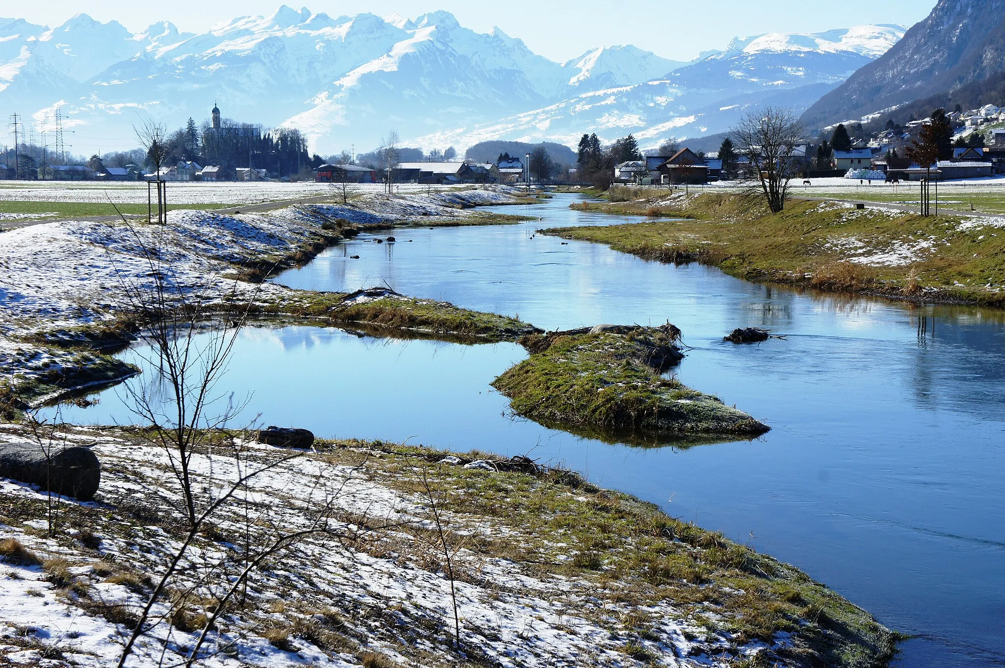 Photo showing: Blick aus Nordosten auf den mittleren Abschnitt des Rheintaler Binnenkanals unterhalb des Dorfes Rüthi. Im Vordergrund der renaturierte Teil des Kanals. In der Bildmitte ist eine Hängebrücke für Fussgänger über den Kanal zu erkennen. Links auf einem kleinen Hügel steht die kath. Pfarrkirche St. Valentin. Im Hintergrund der Alvier, Gamsberg, Gamserrugg und Chäserrugg (von links nach rechts).