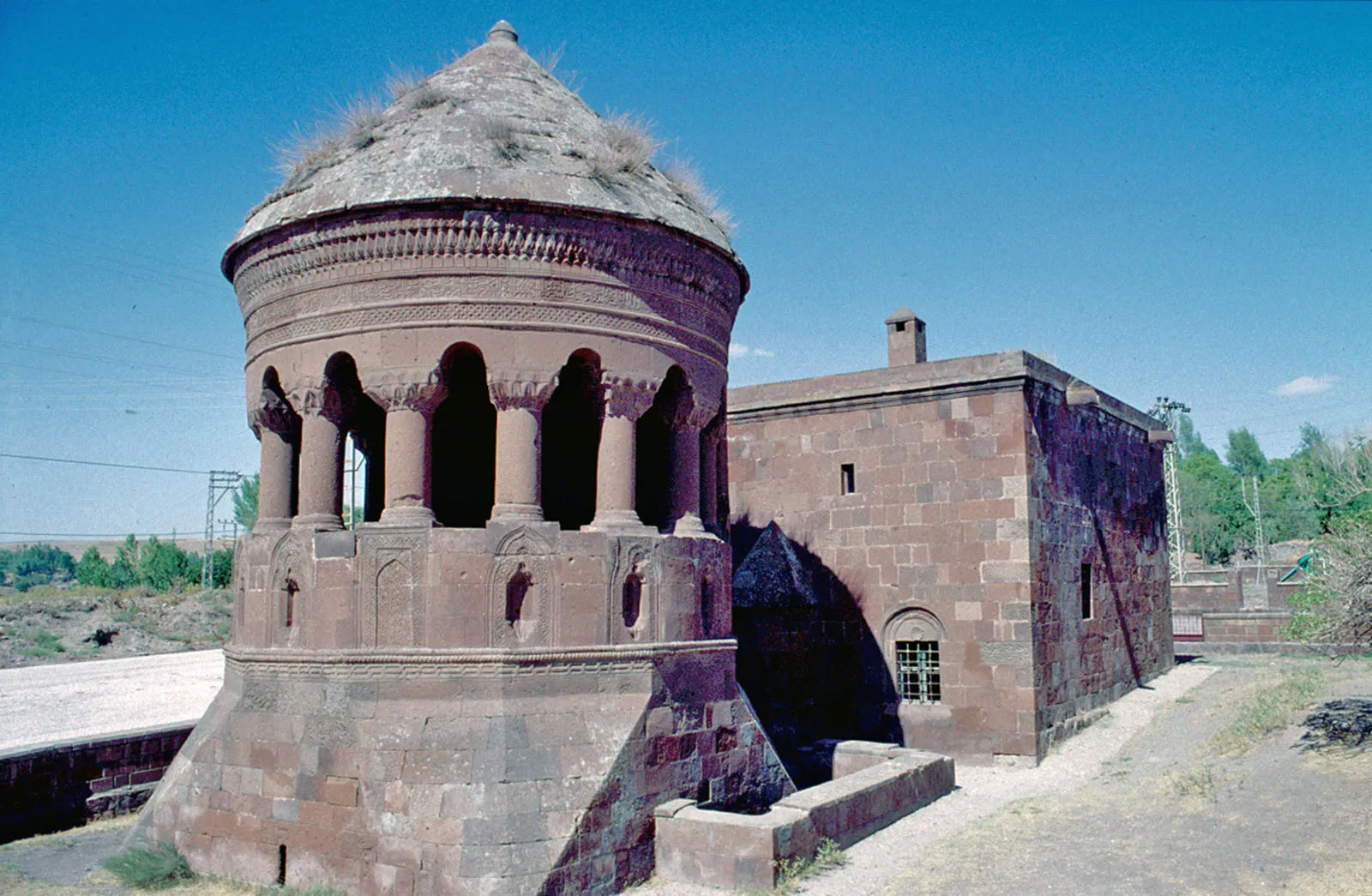 Photo showing: The Emir Bayındır mosque and tomb. It was built in 1491, founded by the governor of a Roha governor, Bayındır. The top floor looks like a round temple with a columnated corridor. The roof is supported by arches that rest on stalactite capitals. Only the back wall that sets it apart from the mosque is completely closed.
This is a detail of the türbe or tomb. Taken from a slide, older than the digital pictures.
Many Seljuk (and later Seljuk-style) mausolea are a stone evocation of the pre-islamic funeral hills of the nomads of Central Asia. During their lives, prominent clan members had their funeral hill (‘kurgan’) prepared; when death came, a circular tent was erected on top of the kurgan, and the deceased’s body was laid out, in order to be greeted a last time by the clan members. After this greeting period, the body was placed in the burial chamber inside the kurgan.

A ‘tent-style’ Seljuk Türbe has two parts: a circular or polygonal room with a pyramidal or cone roof, where a cenotaph sarcophagus can be visited and honoured; this is the part referring to the funeral tent. Beneath this ornamented construction the real burial chamber (‘cenazelik’ or ‘mumyalık’) is to be found, where the deceased’s remains were buried; this is the part referring to the burial hill.