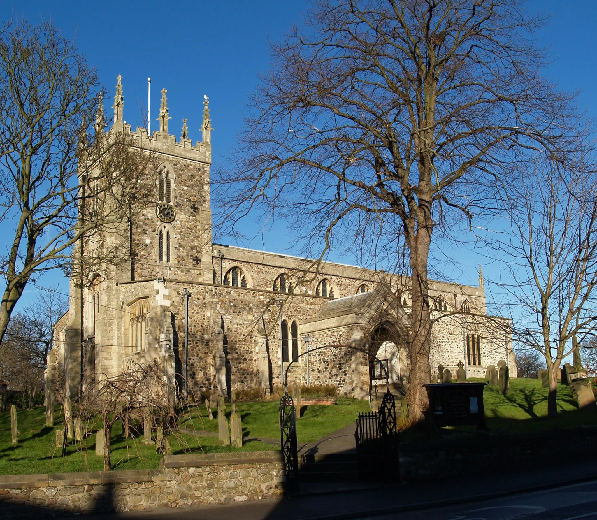 Photo showing: Church of Saint Nicholas, Hornsea, East Riding of Yorkshire, England. Grade I listed building.

"Church. C13 nave and aisles, late C13 tower, altered and enlarged late C14 and C15, restored by Scott 1865-7. Cobbles with freestone dressings. West tower, 4-bay aisled nave with south porch (formerly with south chapel), 4-bay chancel with north and south aisled chapels and eastern crypt. West tower: 3 stages, embraced by westward extension of nave aisles. Chamfered plinth, angle buttresses with offsets, chamfered strings. Pointed openings with Y-tracery under hoodmoulds to 2nd and 3rd stages. Crenellated parapet with eight crocketed pinnacles. Pointed double-chamfered west door under stopped hoodmould. Four-light west window with Perpendicular tracery under 4-centred arch. Nave: 3 buttresses with offsets to westward extension of aisles. 2 grouped lancets to west; similar window to east in blocked pointed chamfered opening to former south chapel. Clerestory: 3-light pointed windows with Perpendicular tracery. Low parapet with moulded coping. Pointed south door under oblong hoodmould with blank trefoils and mouchettes to spandrels. Chancel: moulded plinth, corner buttresses, 3 grouped lancets to south aisle. East bay (added 1430) has five-light pointed windows with Perpendicular tracery to north and south walls. Clerestory: three 3-light pointed window with Perpendicular tracery. Seven-light pointed east window with Perpendicular tracery. Low parapet with crocketed pinnacles and cross finial to coped gable. Interior: triple-chamfered tower arches to north and south. North and south nave arcades of pointed arches with continuous double chamfers: the north on high bases, the south on similar, lower, bases. Pointed chancel arch with continuous fluted mouldings dying into plain bases. Chancel: pointed arches to north and south arcades with continuous chamfer and scrolled hoodmould on high moulded base. Giant blank pointed arches with continuous chamfer enclosing clerestory windows. Crypt, of unknown date, below east bay reached by spiral stair from north choir aisle and small door with shouldered arch (perhaps a former window) beneath east window of chancel. Fireplace with rebated opening under segmental arch to north wall; inserted early C19 double brick vault. Early C13 octagonal font, each face with blank paired lancets in low relief, to nave west end. Memorials: north nave aisle, east end: C13 recumbent effigy in secular clothing. From the church of Saint Giles, Goxhill. Recumbent effigies of a knight and lady from the church of Saint Helena and Saint Mary Magdalene, Nunkeeling; late C13. Chancel south aisle, east end: alabaster tomb chest to Revd. Anthony St. Quintin, died 1430. Sides with blank shields in quatrefoils, remains of inscription to edge of covering slab. This is covered in early graffiti, including large numbers of 'footprints'." Church of Saint Nicholas, Hornsea