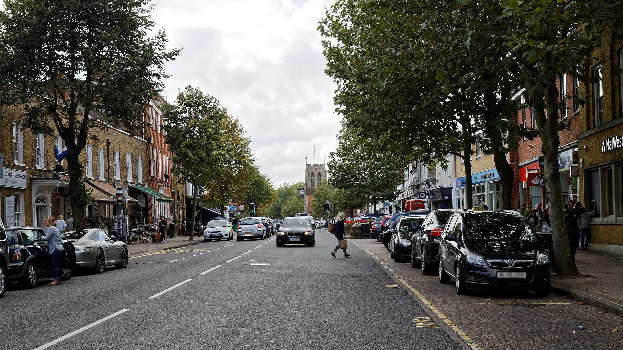 Photo showing: Looking south-west on Epping High Street (the B1393 road), towards St John the Baptist Church, in Epping, Essex, England. Software: RAW file lens-corrected, optimized and converted to JPEG with DxO OpticsPro 10 Elite, and likely further optimized and/or cropped and/or spun with Adobe Photoshop CS2.