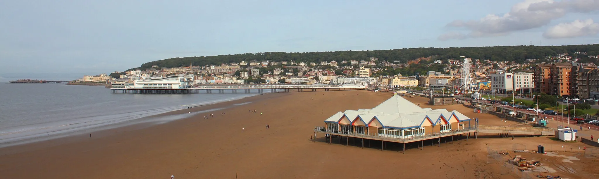 Photo showing: The view across Weston-super-Mare from a viewpoint above the Tropicana. The Birnbeck (or 'Old') pier juts out from the end of Worlebury Hill. The Grand Pier hides Knightsone Island and the north end of the sea front. The former Sea Life Centre is the third pier in the foreground, currently used as a café.