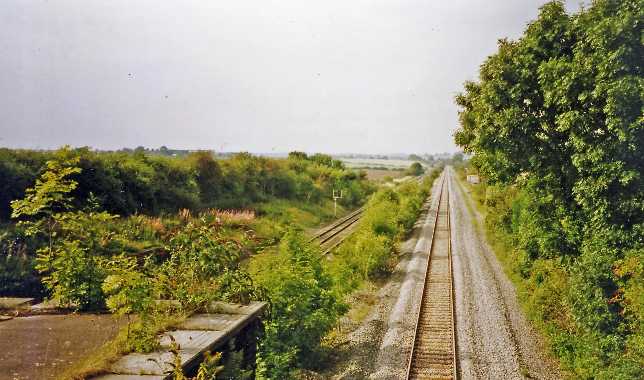 Photo showing: Site of former Irchester station.
View NW from the B569 bridge, towards Wellingborough, Kettering and the North: ex-Midland Main Line from London St Pancras. The single line on the right is the Slow line, formerly double-track, which had been built in 1880-84 with a tunnel under Souldrop Summit for goods trains to avoid the steep gradients of the original route; it was singled in 1987. Irchester station was closed to passengers 7/3/60, to goods 4/1/65.