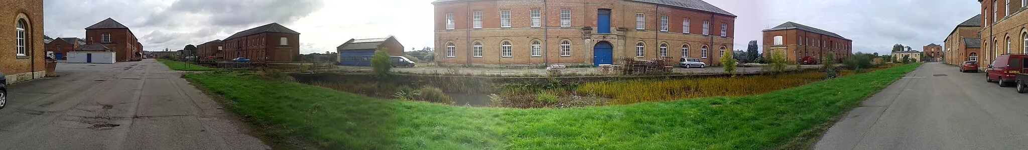 Photo showing: Former Ordnance Depot at Weedon Bec. The extensive barracks were begun in 1803 during the Napoleonic wars, but most of the buildings (including 12 powder magazines) have been demolished. This is a 180-degree panoramic shot and therefore the straight road appears as two roads. In fact it is parallel to the grass verge and canal in the foreground.