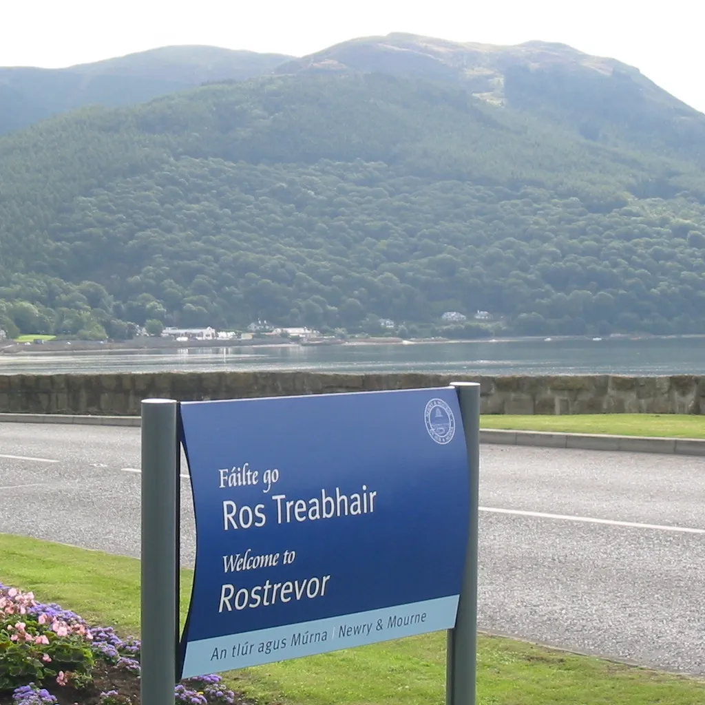 Photo showing: Welcome sign in Rostrevor in English and Irish (with Rostrevor Forest in the background)