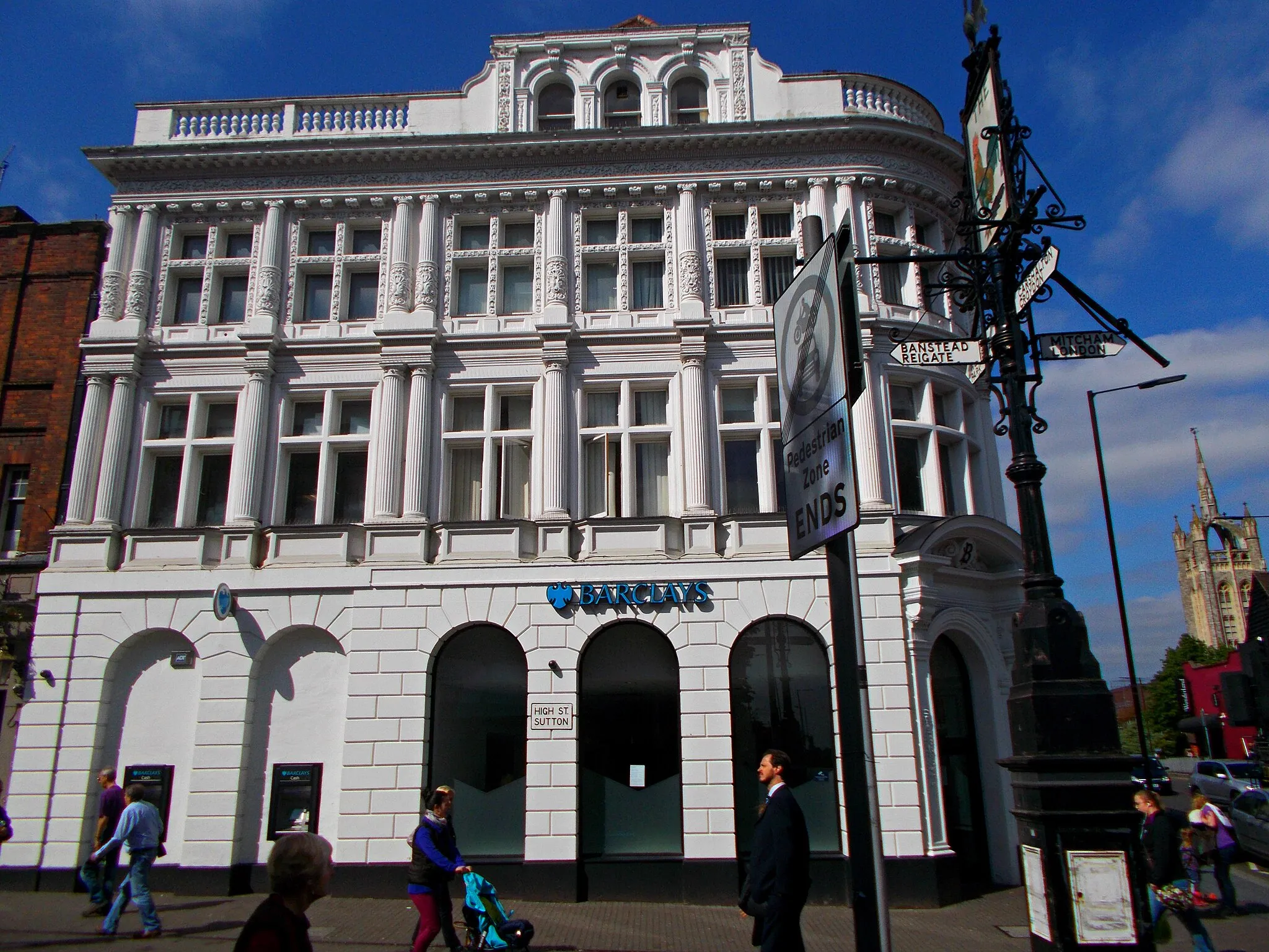 Photo showing: The Barclays Bank building is a four-storey locally listed building overlooking the historic crossroads in the town centre of Sutton in the south London Borough of Sutton. It was built in 1894, originally as the London and Provincial Bank.
The building is situated at the junction of Cheam Road and Sutton High Street, the location of the crossroads of the historic turnpike roads which passed through the town. It is located in the Sutton Town Centre High Street Crossroads Conservation Area, which was designated on 9 May 2011.
The building is four storeys in height and of ornate design. It forms a prominent landmark when arriving in the town centre from a westerly direction. There is a series of arches at ground level within rusticated stonework. The main entrance is on the corner where the two roads meet, rounded in shape and surrounded by an ornate architrave and segmental pediment, which together serve to create a sense of importance for the bank.

The second storey has fluted columns and simple cross windows. The windows, columns and dentil on the third storey are surrounded by decorative carvings. The attic is on the fourth storey and set within a decorative, balustraded parapet under which there is a decorative frieze.