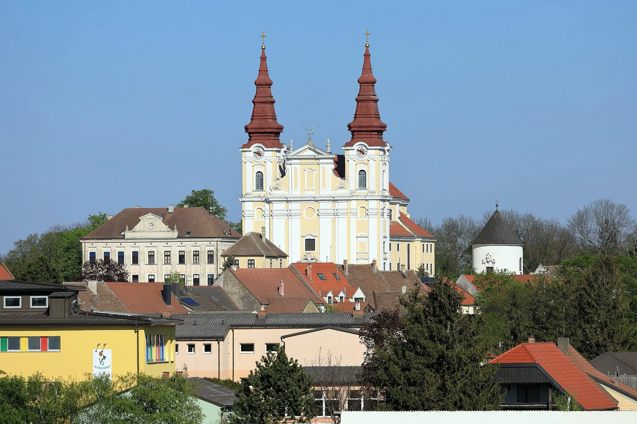 Photo showing: Westansicht des Zentrums der niederösterreichischen Marktgemeinde Wullersdorf. Im Bildzentrum die ortsbildbeherrschende Pfarrkirche hl. Georg. Links davor die ehemalige Schule, ein dreigeschoßiges Gebäude mit Walmdach sowie Giebelaufsatz und rechts der Rundkarner.