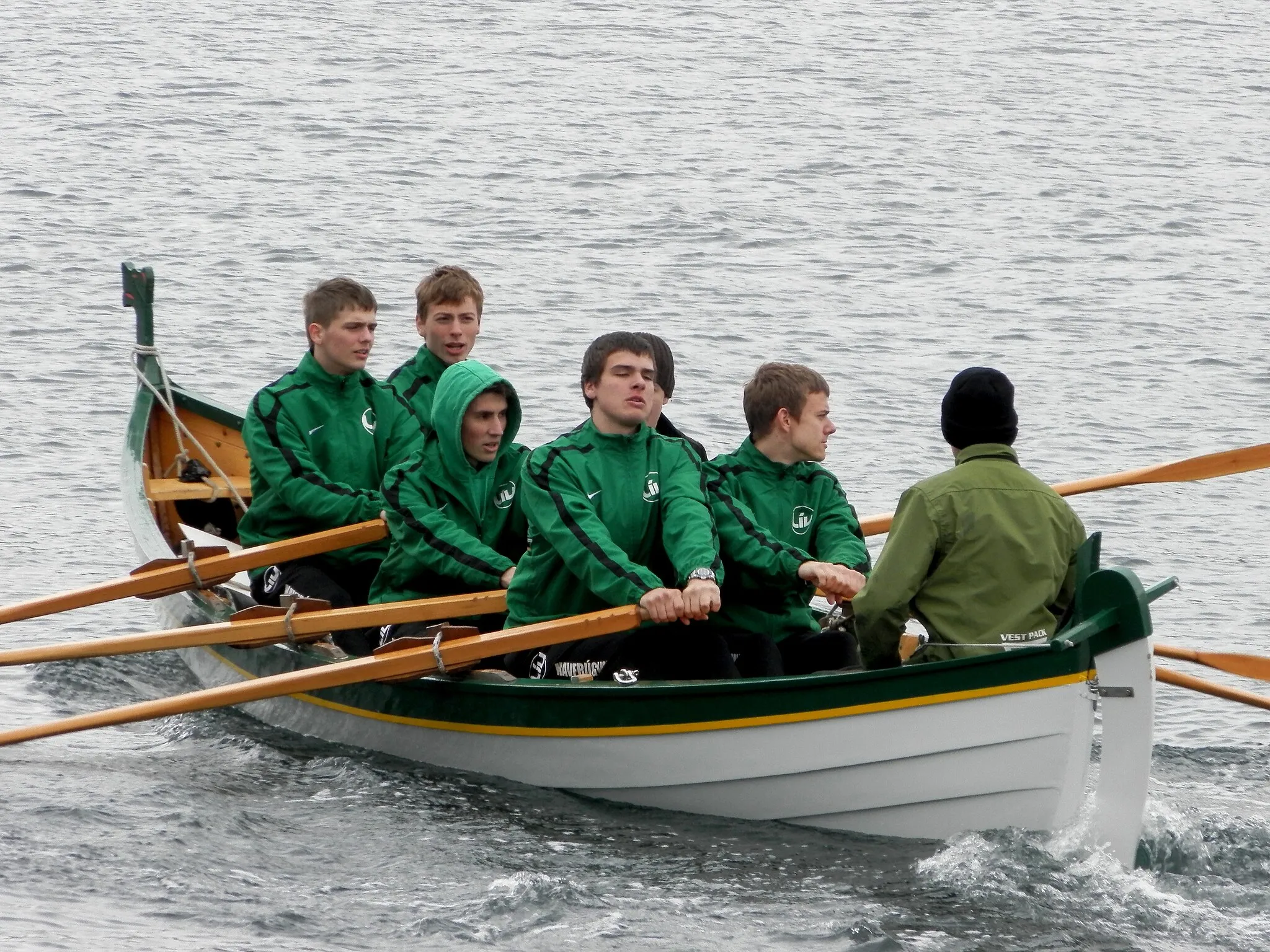 Photo showing: Havfrúgvin is a Faroese wooden rowing boat from the village Vestmanna, it is a 5-mannafar. This photo was taken on Eystanstevna in Runavík, Faroe Islands. They are on their way out to the start here. The rowing competitions in the Faroe Islands are on the sea, in fjords or sounds. There are 7 competitions in June and July. There are winners for every competition and there are final winners of the the races, they will be Faroese Champions. There are 9 categories in the Faroese Championship: Children, boys under 15, in 5-mannafør, children, girls under 15, in 5-mannafør, 5-mannafør boys under 18, 5-mannafør girls under 18, 5-mannafør women, 6-mannafør men, 6-mannafør women, 8-mannafør men and 10-mannafør men. A 5-mannafar is the smallest boat type, it is for 6 rowers, and a 10-mannafar is the largest boat type, for 10 rowers. One of the rowers in this boat, Torstein Brattaberg, is also competing in indoor rowing. He won the Danish Junior Championship in 2011 and 2012 and the Nordic Junior Championship in 2000 meter indoor rowing for boys 15-16.