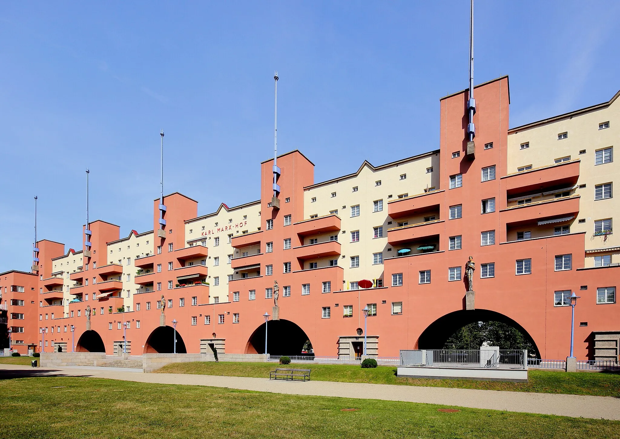 Photo showing: Der stark zurückversetzte Mitteltrakt mit seinen sechs monumentalen Türmen der denkmalgeschützten Wohnhausanlage Karl-Marx-Hof der Gemeinde Wien im 19. Wiener Gemeindebezirk Döbling. Der Bau mit 1382 (aktuell rd. 1270) Wohneinheiten wurde von 1927 bis 1933 nach Plänen des Architekten Karl Ehn errichtet (die offizielle Eröffnung der Anlage fand am 12. Oktober 1930 statt). Er ist wohl der repräsentativste, sicherlich aber der bekannteste kommunale Wohnbau der Stadt Wien als Musterbeispiel eines monumentalen Superblocks. Er erstreckt sich über 1.100 Meter entlang der Heiligenstädter Straße und ist damit der längste zusammenhängende Wohnbau der Welt. Die vier Figuren über den Rundbögen sind Werke des Bildhauers Josef Franz Riedl (1884–1965) und symbolisieren die „Freiheit“, die „Fürsorge“, die „Aufklärung“ und die „Körperkultur“.