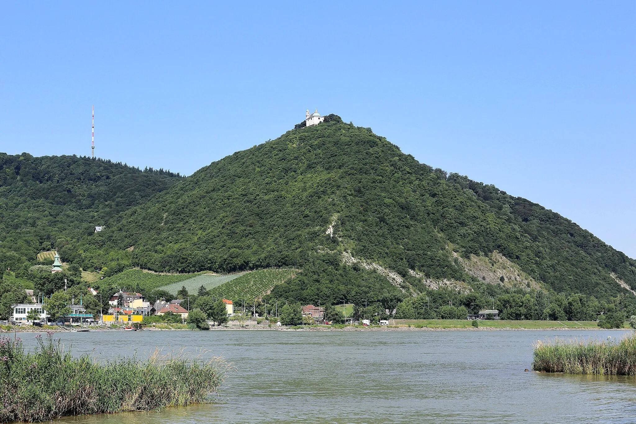 Photo showing: Südostansicht des 425 Meter hohen Leopoldsberges im 19. Wiener Gemeindebezirk Döbling mit der Leopoldsbergkirche. Im Vordergrund die Donau, links der Ort Kahlenbergerdorf und dahinter der 484 Meter hohen Kahlenberg mit dem Sender Kahlenberg.