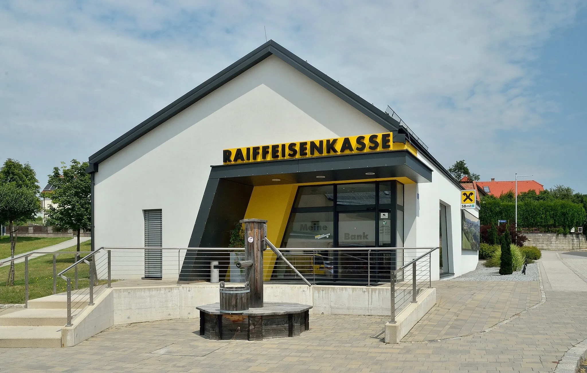 Photo showing: Raiffeisenkasse Hochwolkersdorf mit einem Brunnen davor, Gemeinde Hochwolkersdorf, NÖ.