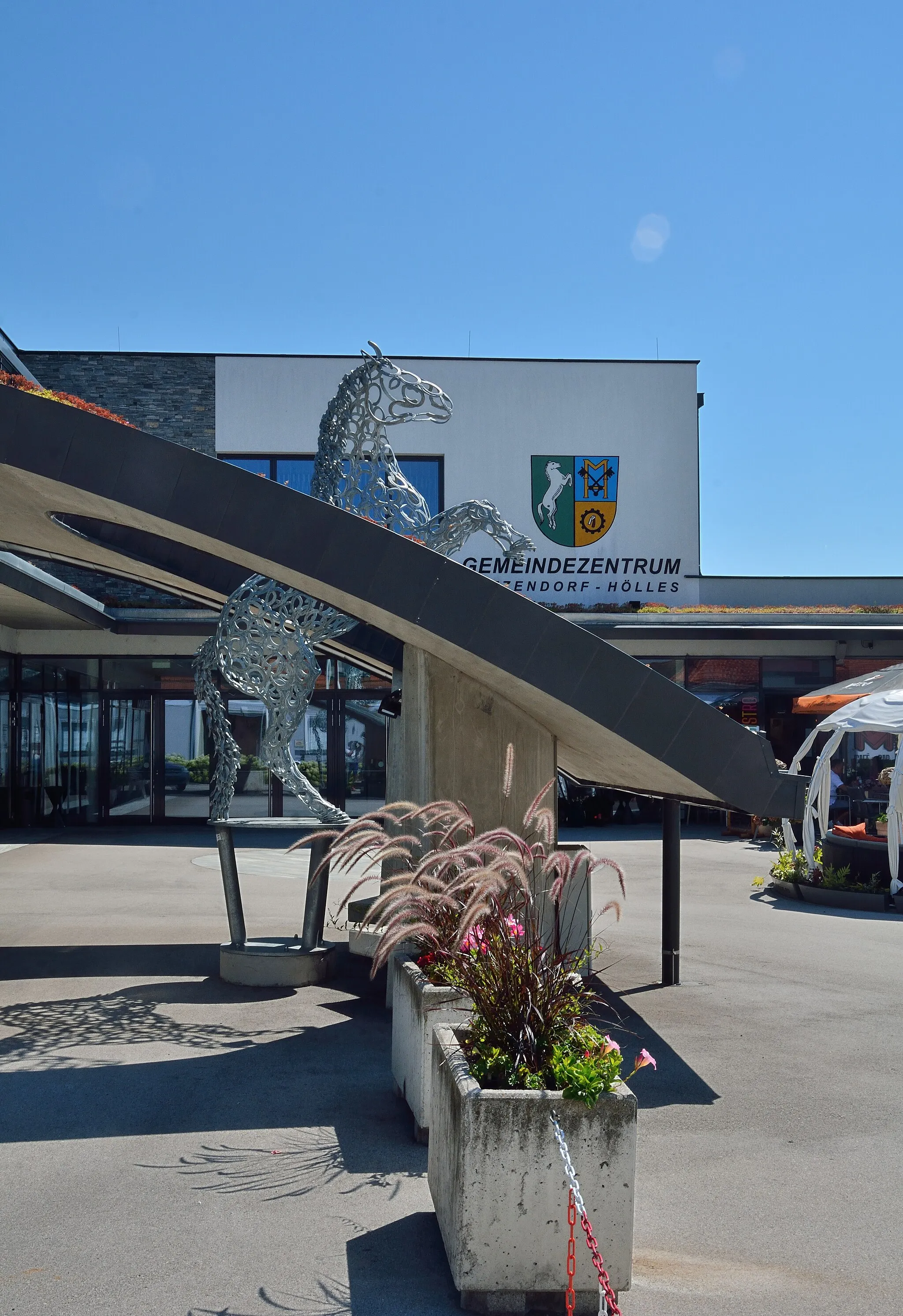 Photo showing: Das Gemeindeamt Matzendorf-Hölles in der Badner Straße steckt mit der Raiffeisen-Bank unter einem Dach. Vor dem Gebäude eine Pferdeskulptur aus Hufeisen, die dem Pferd im Wappen der Gemeinde entspricht. Hier in der Gegenüberstellung mit dem Wappen deutlich zu sehen.