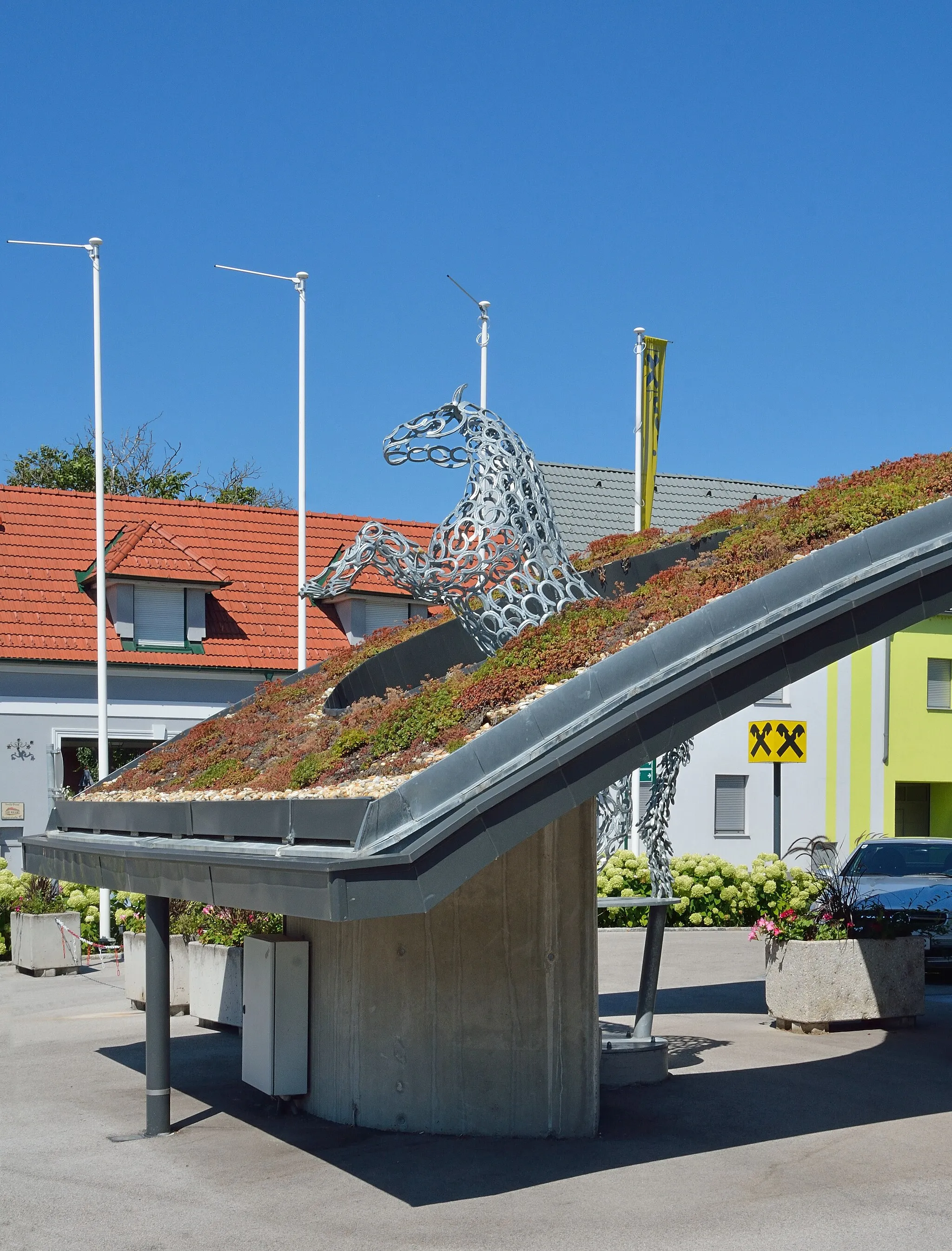 Photo showing: Das Gemeindeamt Matzendorf-Hölles in der Badner Straße steckt mit der Raiffeisen-Bank unter einem Dach. Vor dem Gebäude eine Pferdeskulptur aus Hufeisen, die dem Pferd im Wappen der Gemeinde entspricht. Auf der anderen Seite entspricht das Pferd auch der Hälfte des Giebelkreuzes von Raiffeisen.