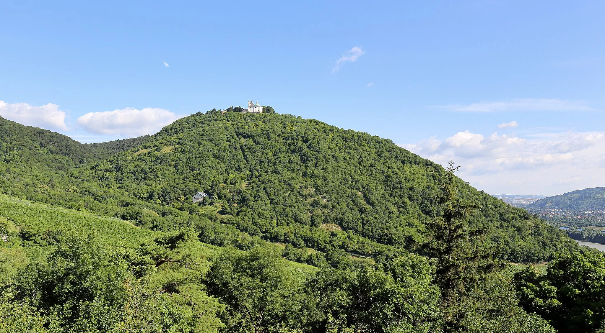 Photo showing: Südsüdostansicht des 425 Meter hohen Leopoldsberges im 19. Wiener Gemeindebezirk Döbling mit der Leopoldsbergkirche, dem Heimkehrer-Gedächtnismal und der Leopoldsbergburg. Links der Anstieg des 484 Meter hohen Kahlenberges und rechts von vorne nach hinten: Die Donau, die Donauinsel, das Entlastungsgerinne Neue Donau, die Marktgemeinde Langenzersdorf und der 385 Meter hohe Bisamberg.