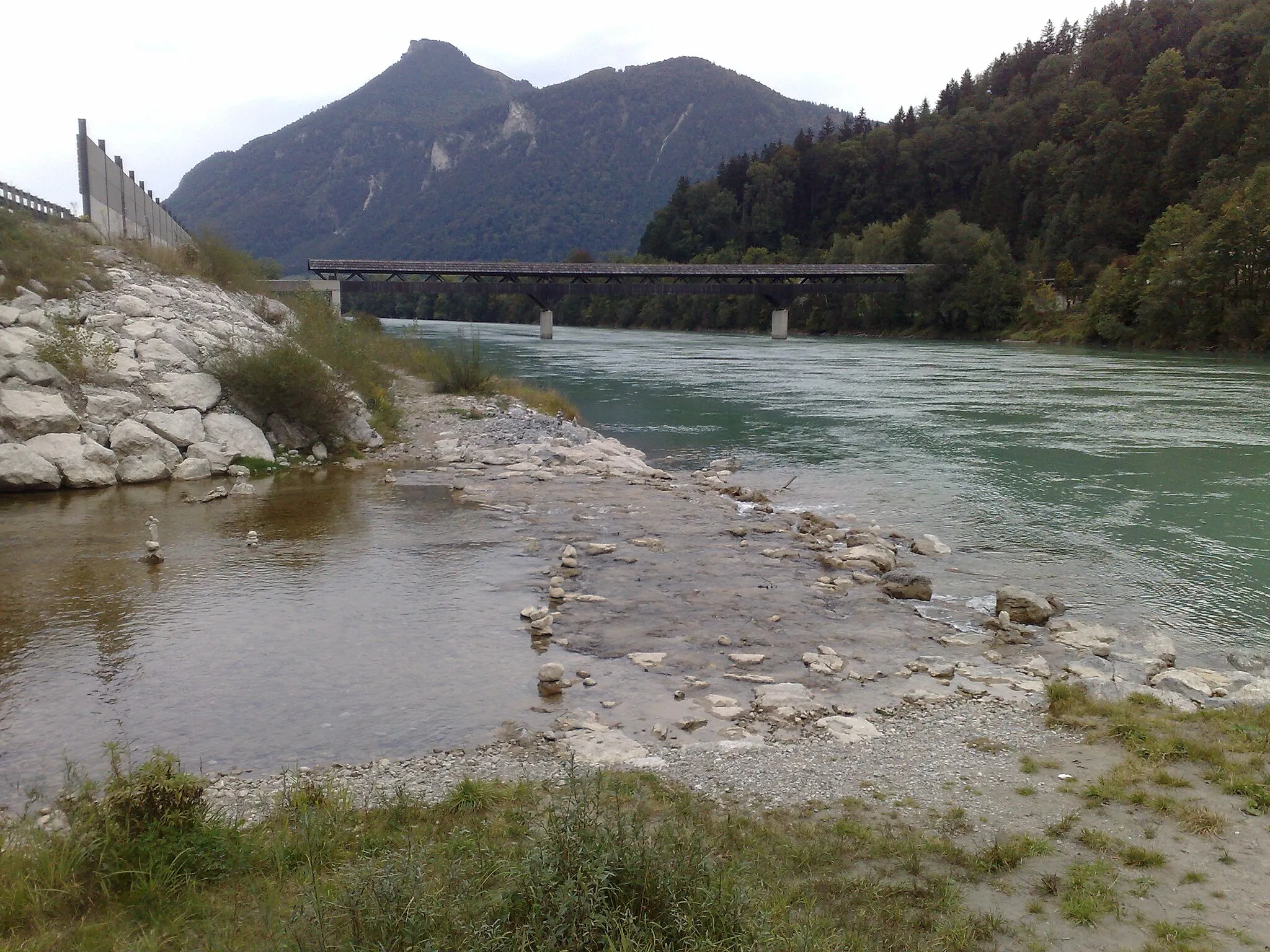 Photo showing: Confluence of the clear creek Auerbach from left (west) here into the north-flowing green-colored river Inn. View towards North. Over the Inn a wooden bridge to Erl-Zollhaus in Tyrol. In the background the Kranzhorn, on whose summit the German-Austrian border runs.